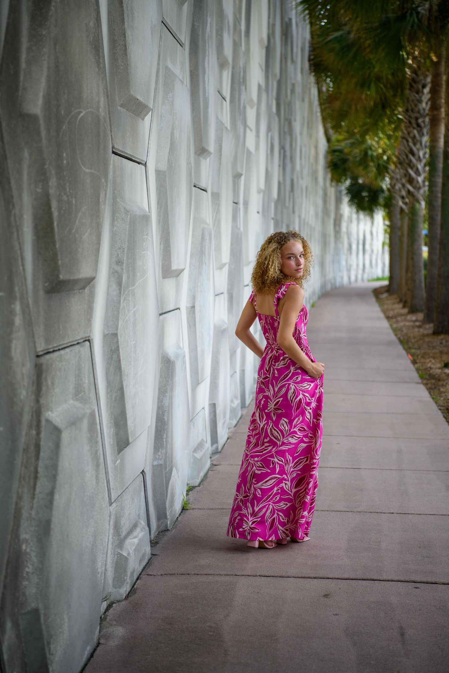 Person in pink floral maxi dress poses along white architectural wall.