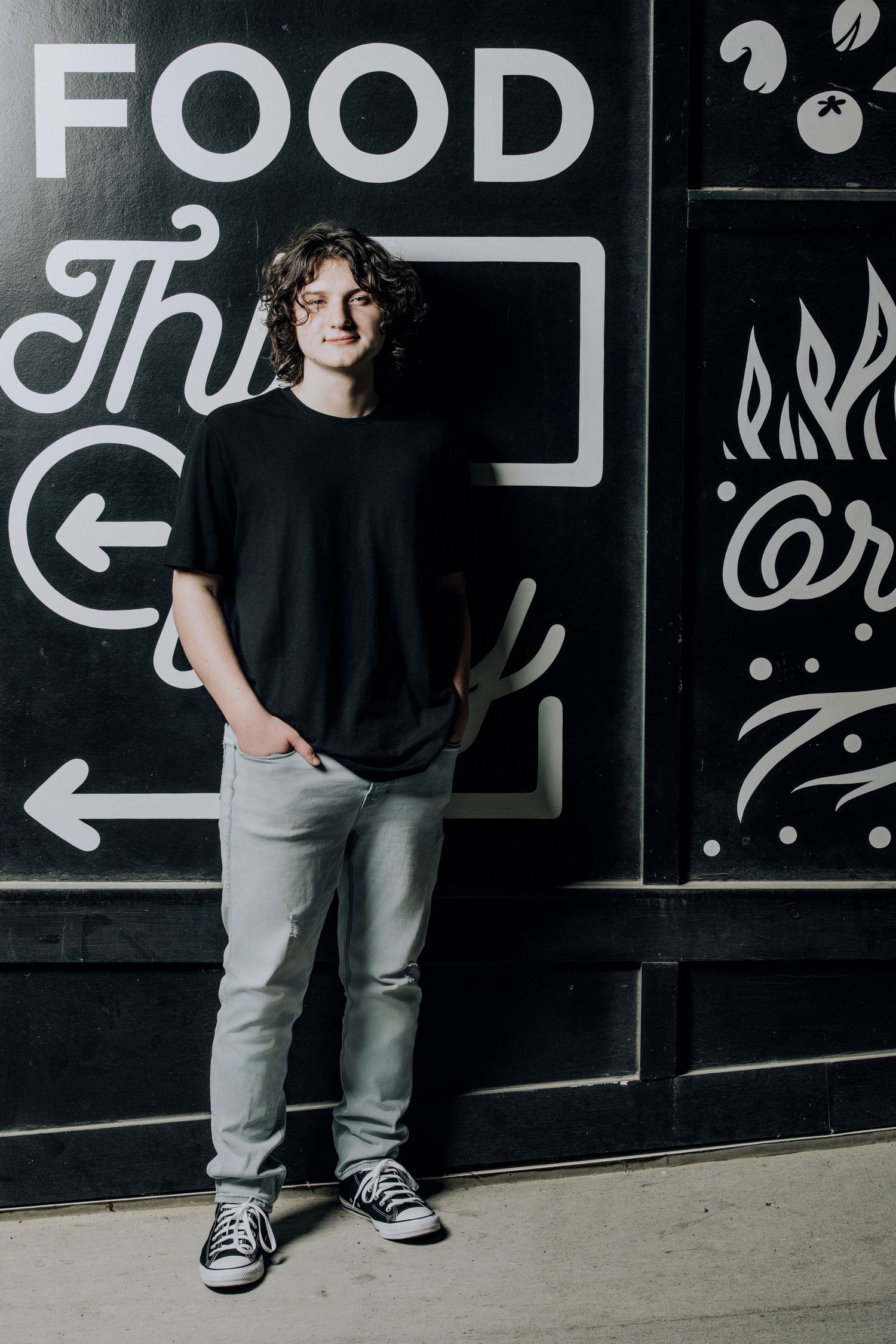 Casual portrait of person in black t-shirt standing against wall with food signage.