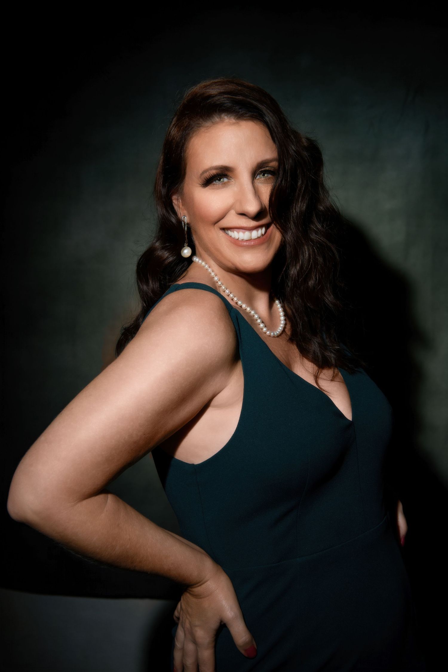 Elegant portrait of a woman in a navy dress wearing pearl jewelry against a dark green background.
