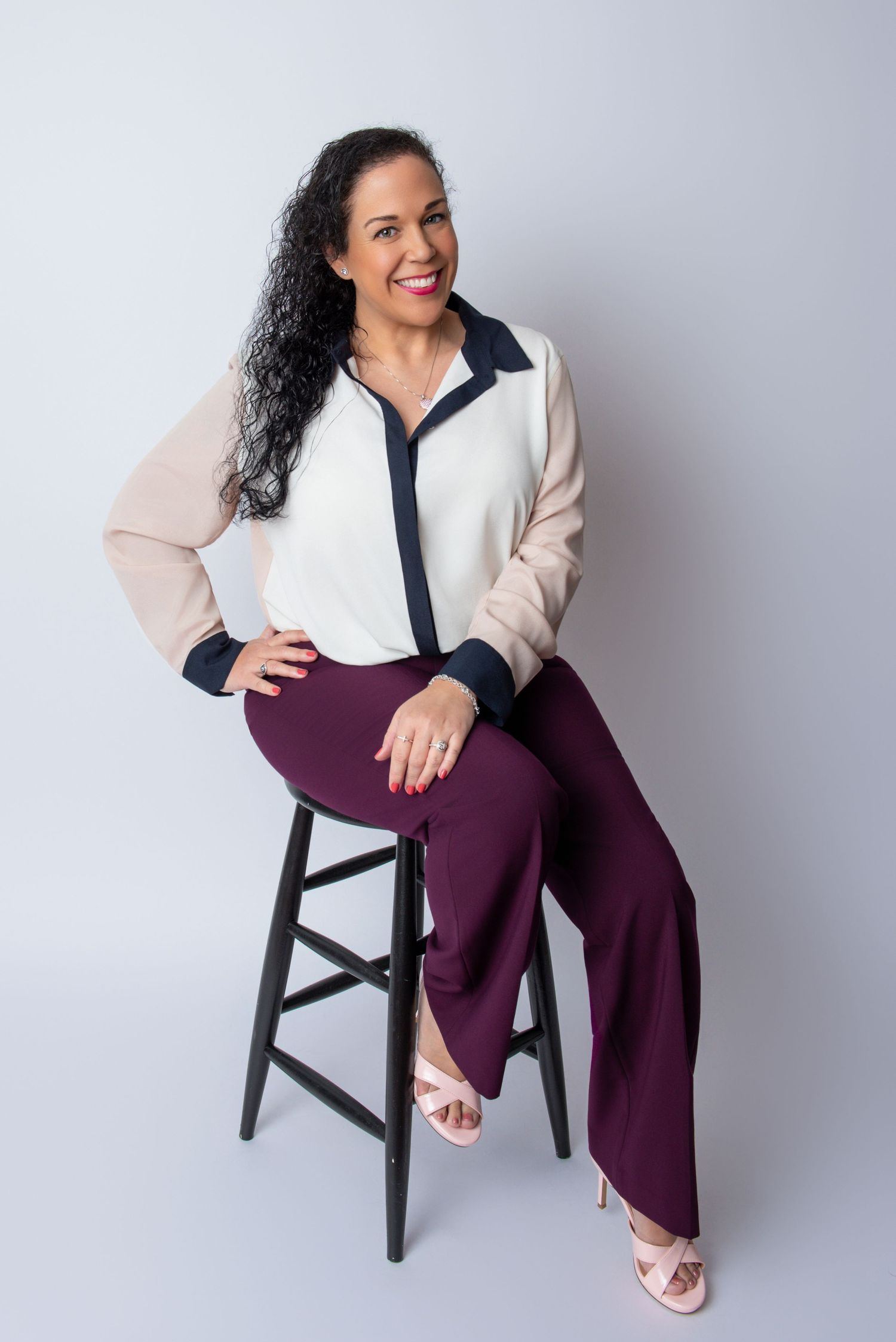 Professional portrait of a woman in business attire sitting on a black stool against a light background.