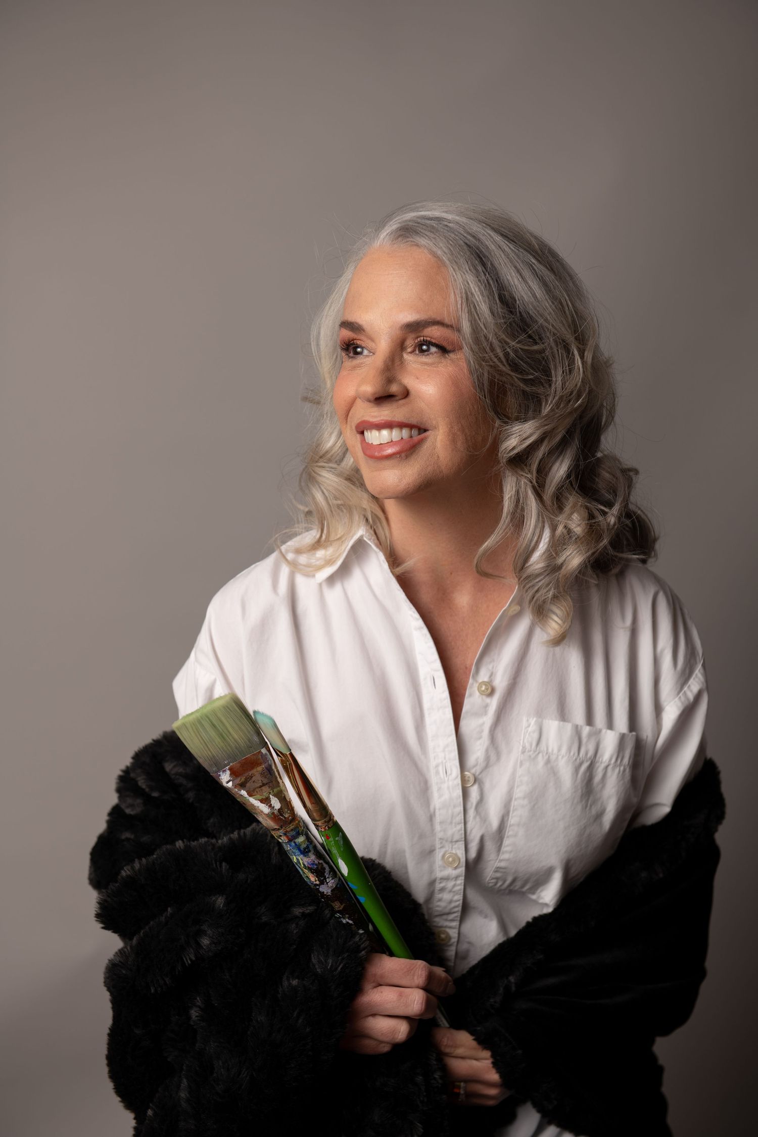 Studio portrait of someone in a white shirt and black fur coat holding paint brushes.