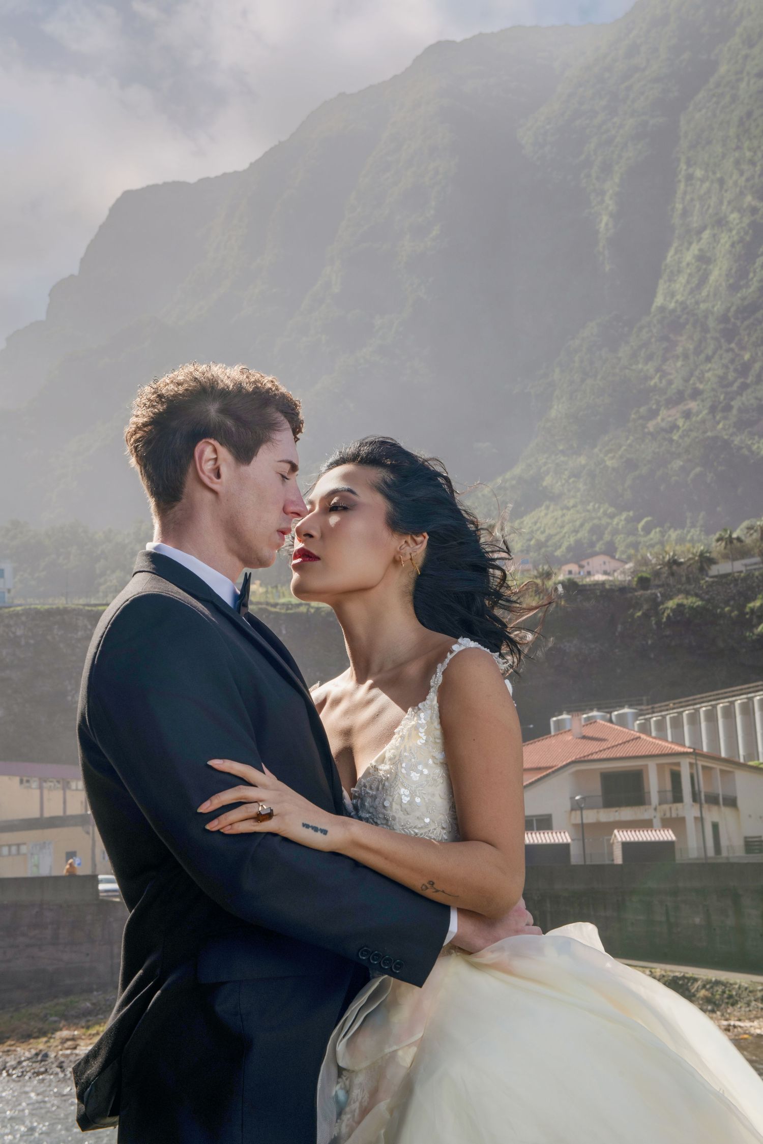 Newlyweds share romantic kiss with dramatic mountain landscape in background.