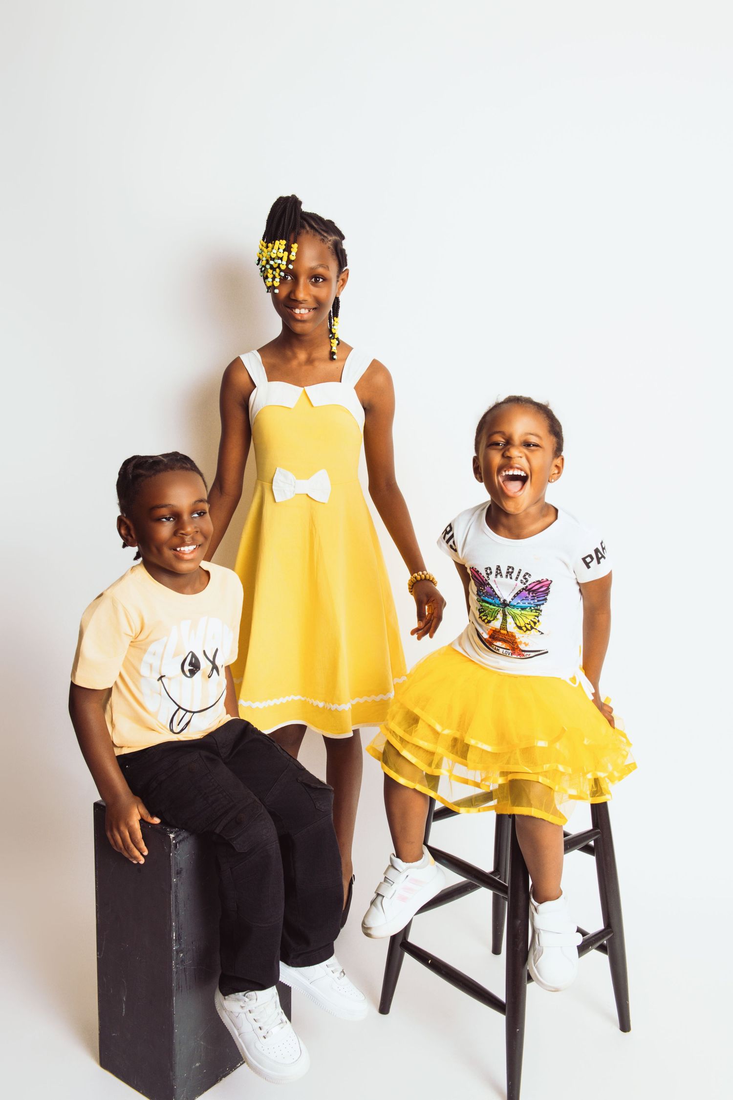 Children in yellow and white outfits sitting on black stools against white backdrop.