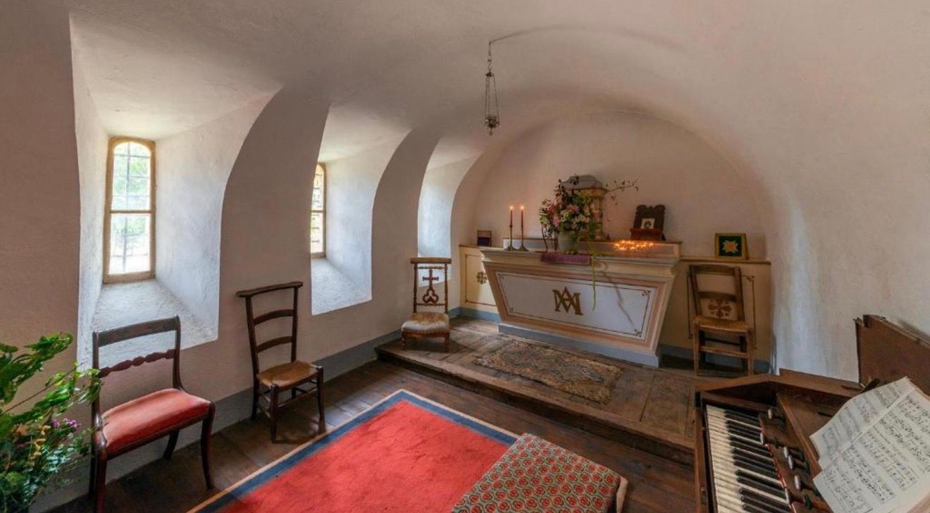 Rustic vaulted stone room with arched ceilings, wooden chairs, red rugs, and an antique altar table with decorative items.