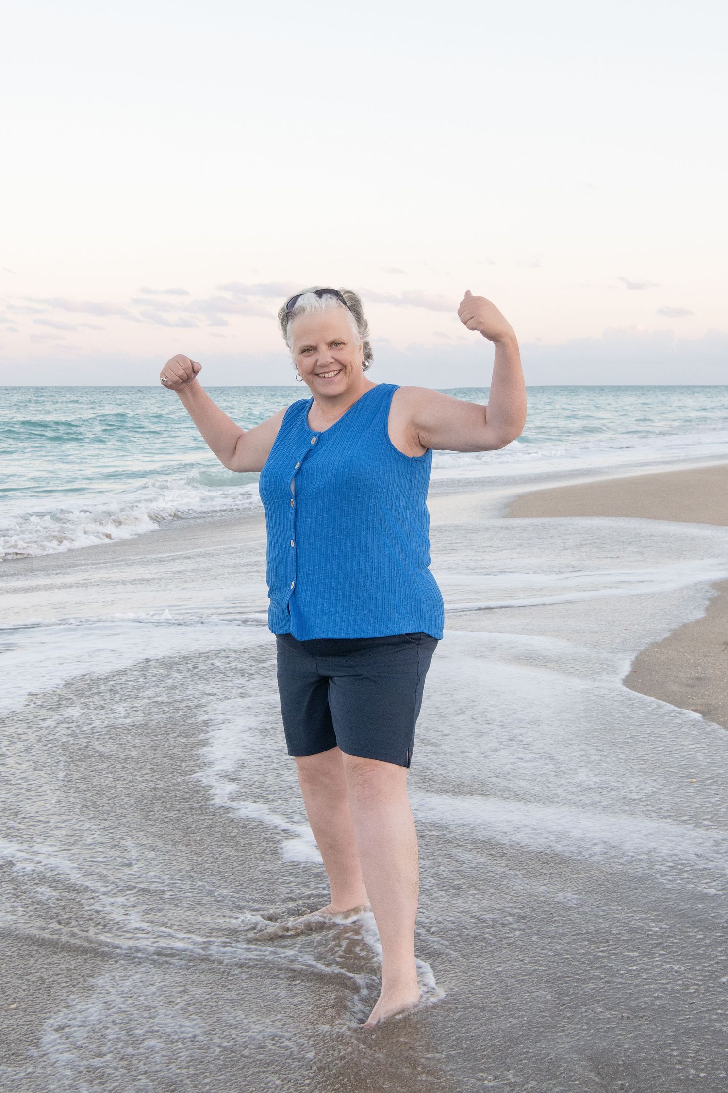 Joyful 50 year old woman in blue tank top celebrating on beach at sunset.