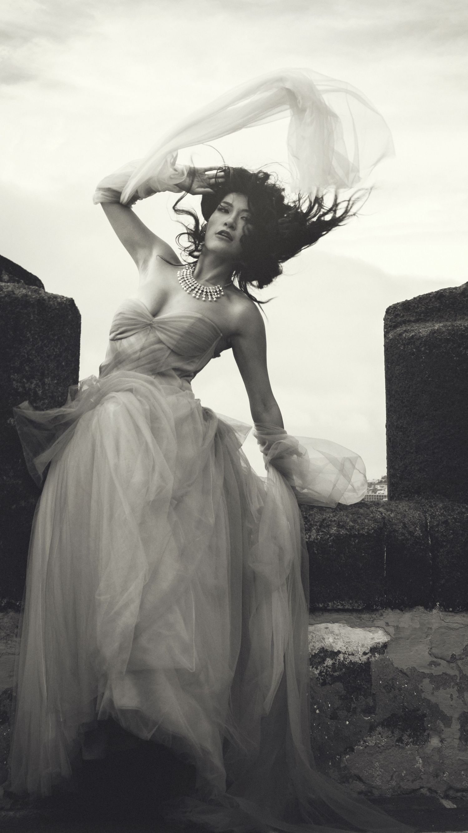 Dynamic black and white fashion photo with flowing dress against stone architecture.