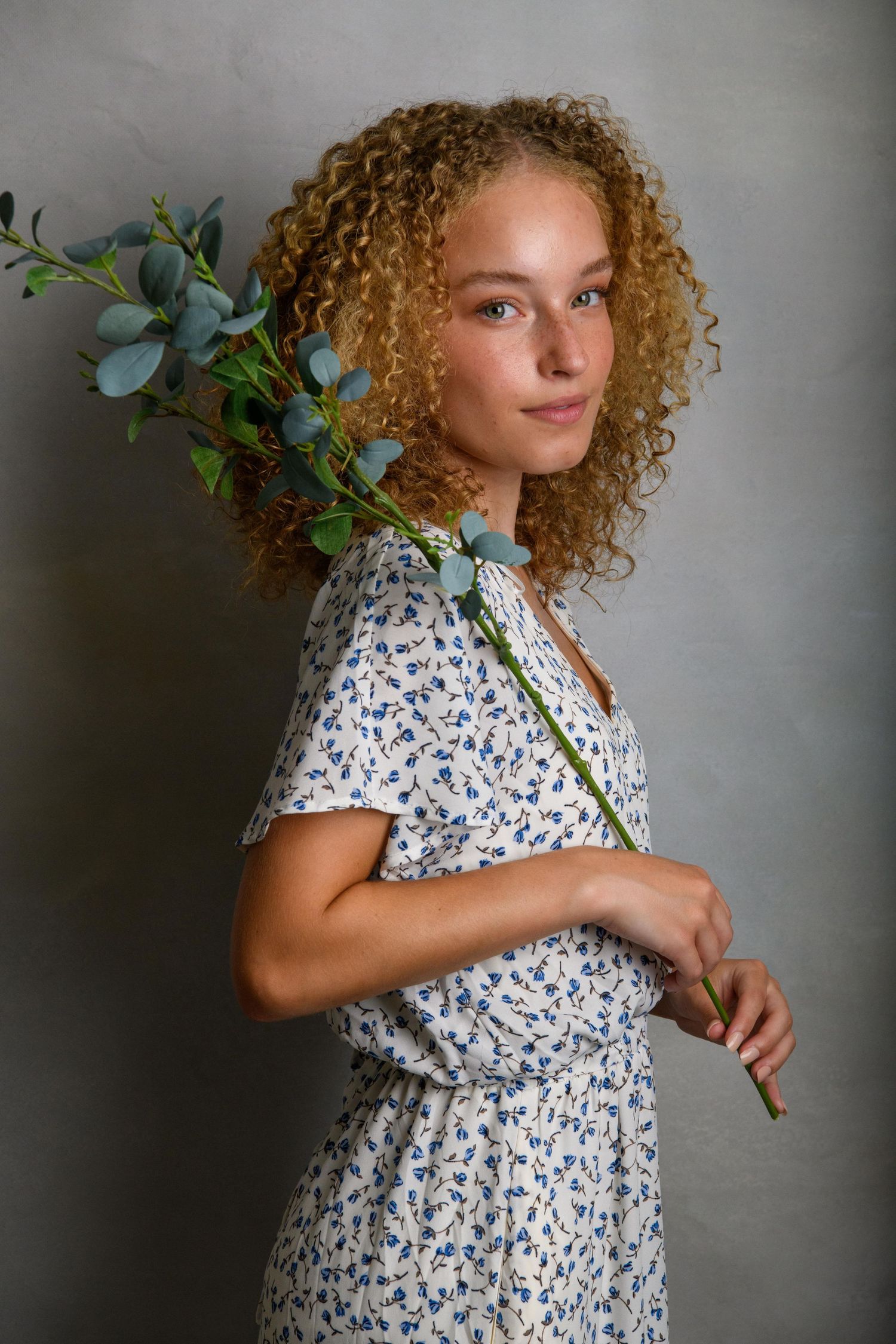 Artistic portrait with curly hair and eucalyptus branch against grey studio backdrop.