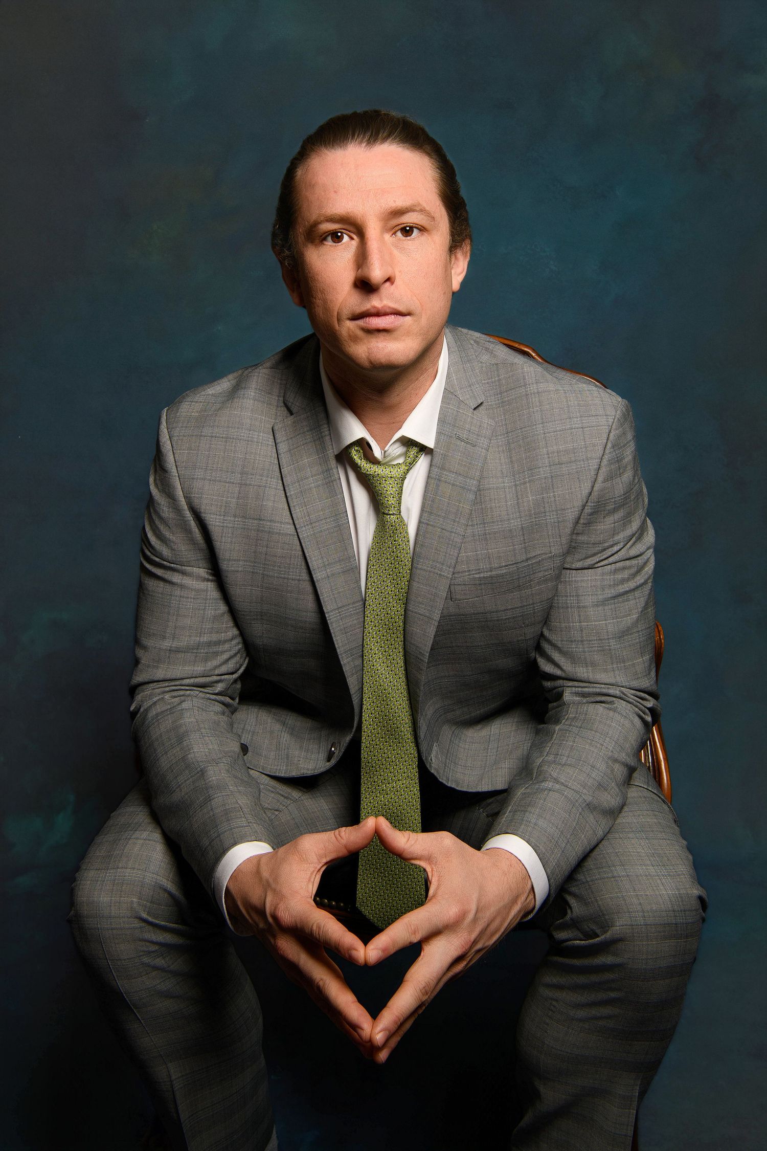 Man in gray suit and green tie sits confidently with hands clasped against a dark blue studio background.