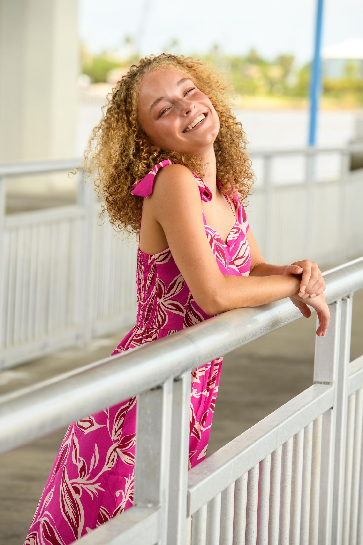 Person with curly hair leans against white railing wearing vibrant pink floral dress.