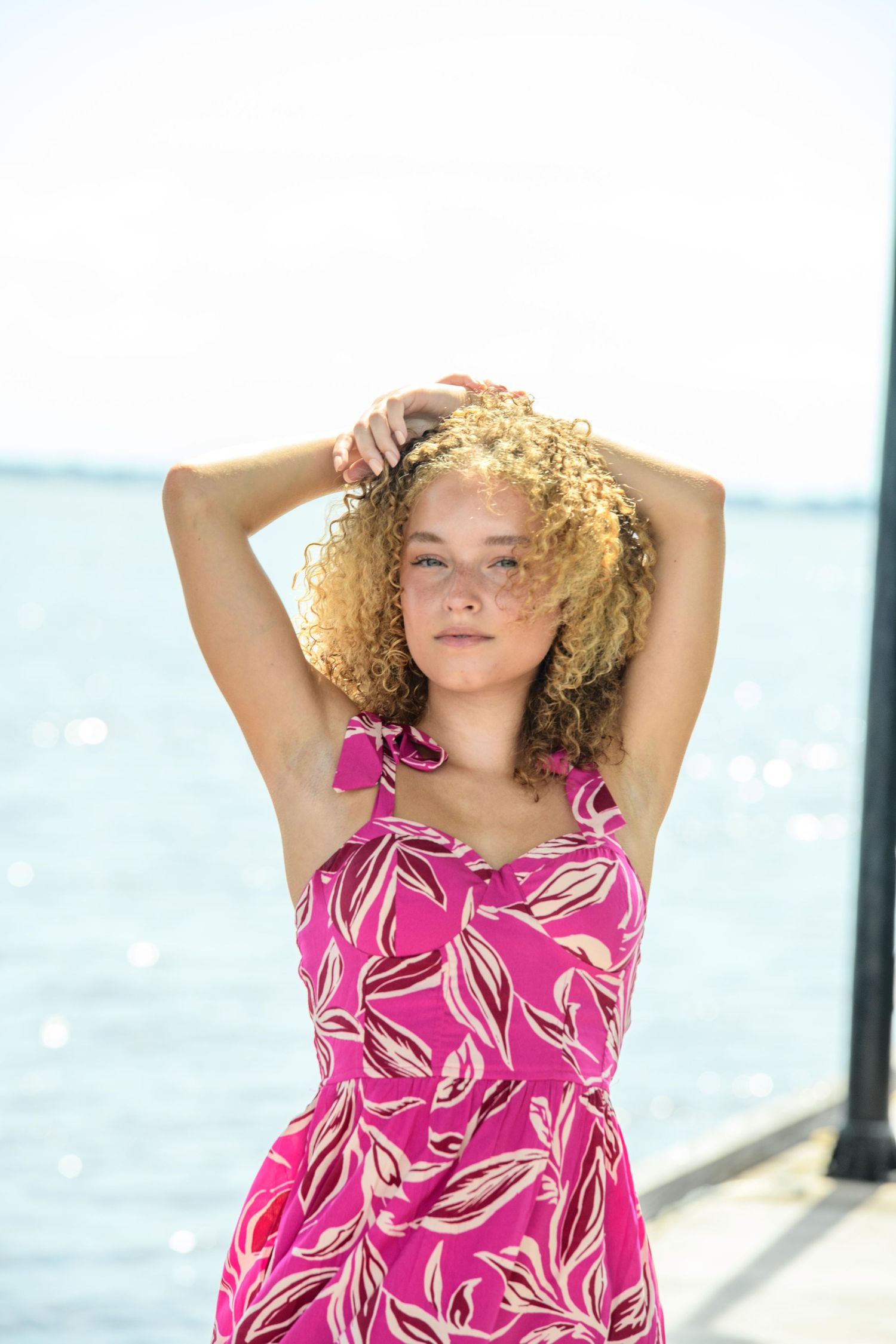 Beachy portrait with curly hair in pink floral sundress against ocean background.