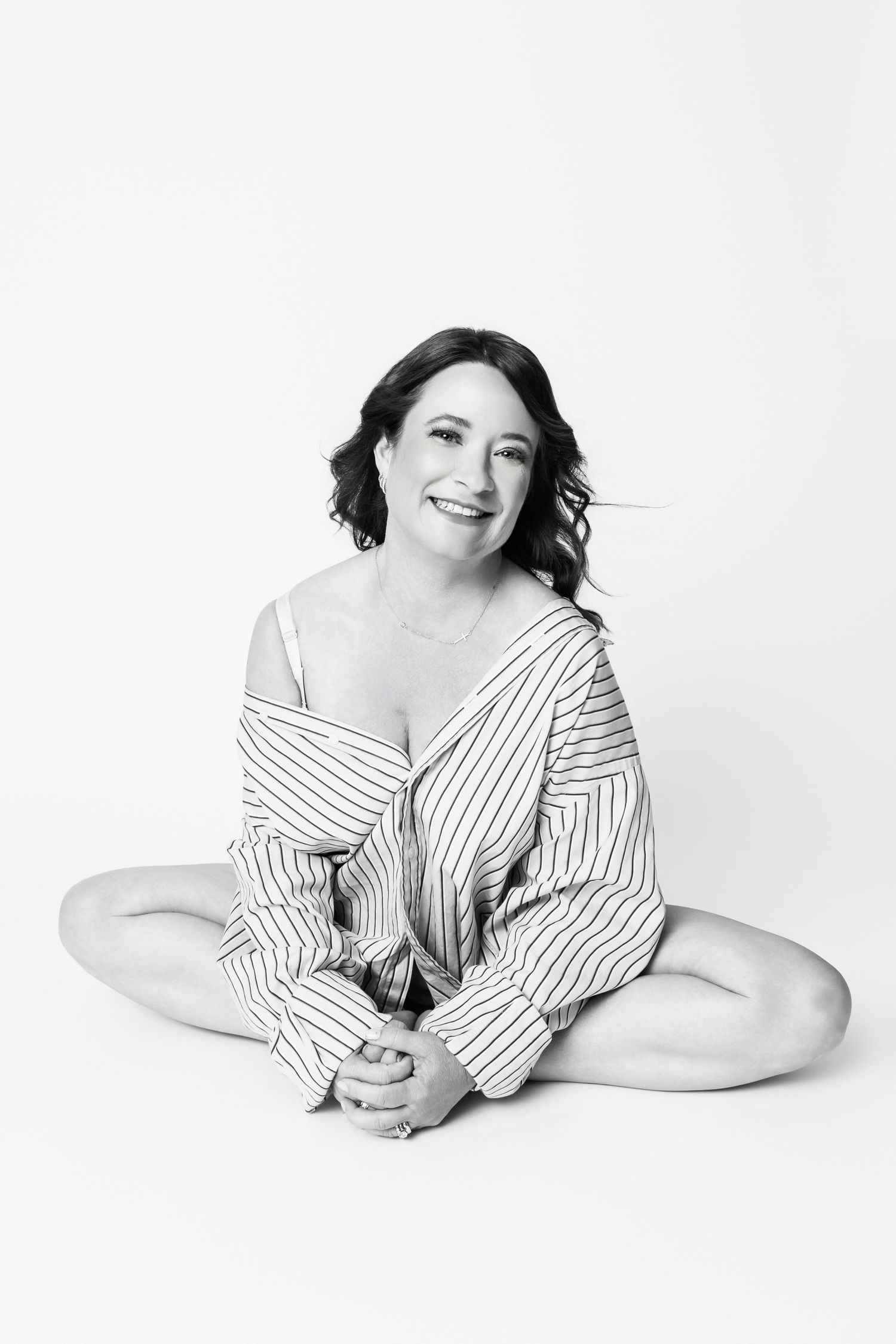 Casual studio portrait in striped button-down shirt sitting cross-legged on white background.
