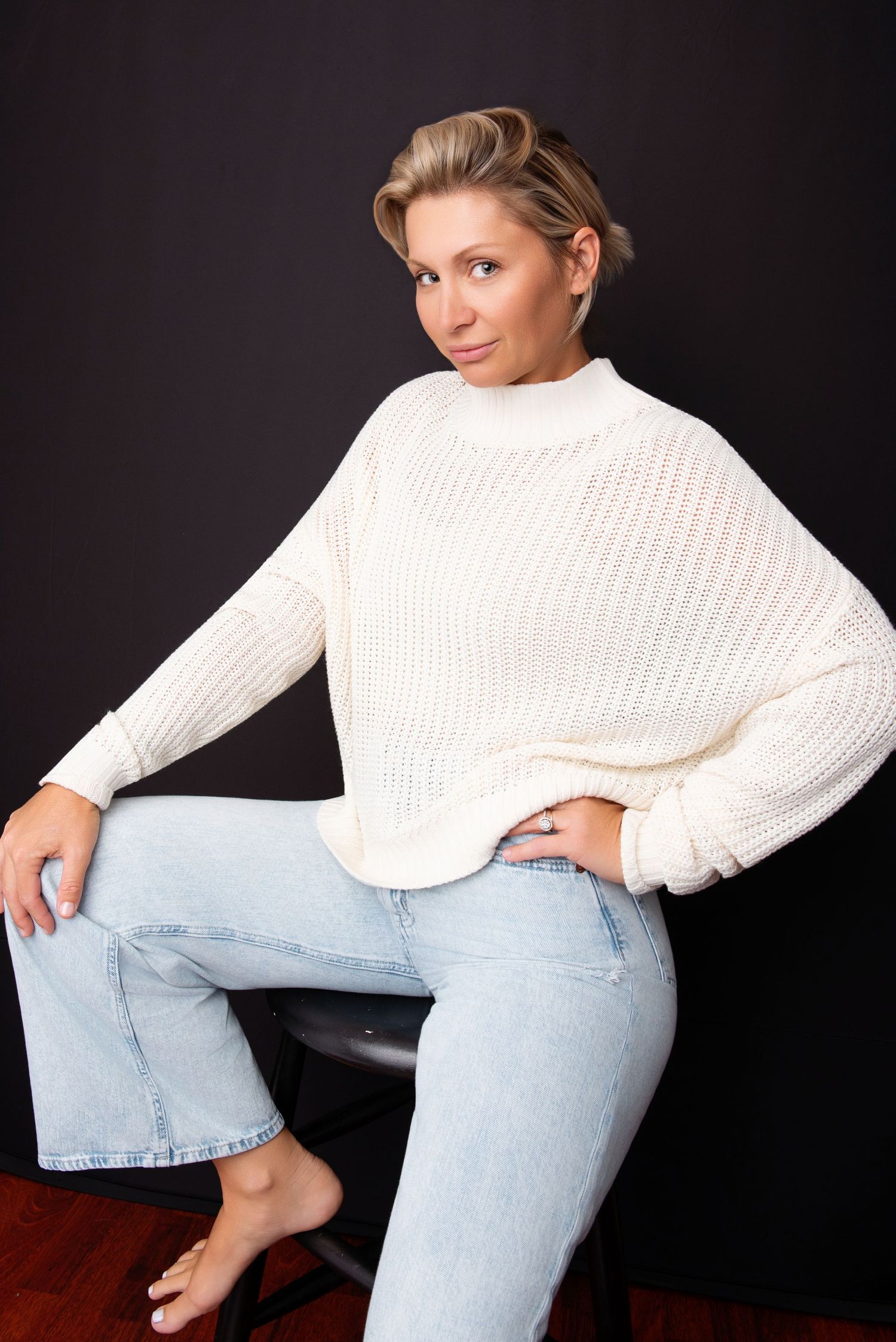 Casual portrait in white sweater and light denim with relaxed pose against dark background.