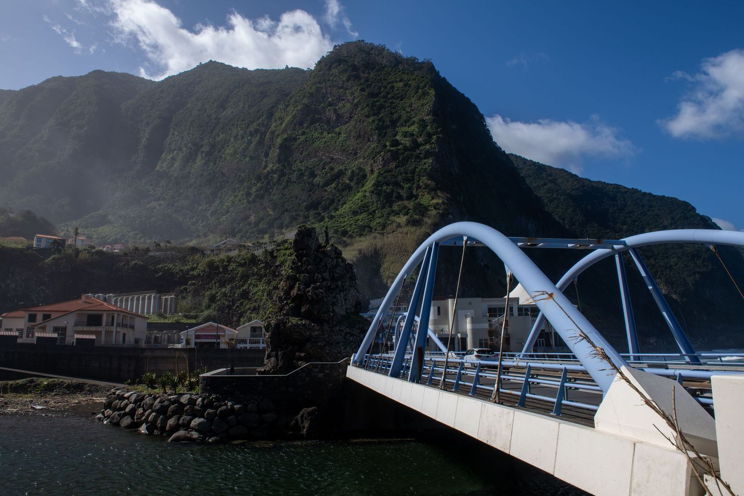 Modern white bridge spans across a coastal inlet with mountains and buildings in the background.