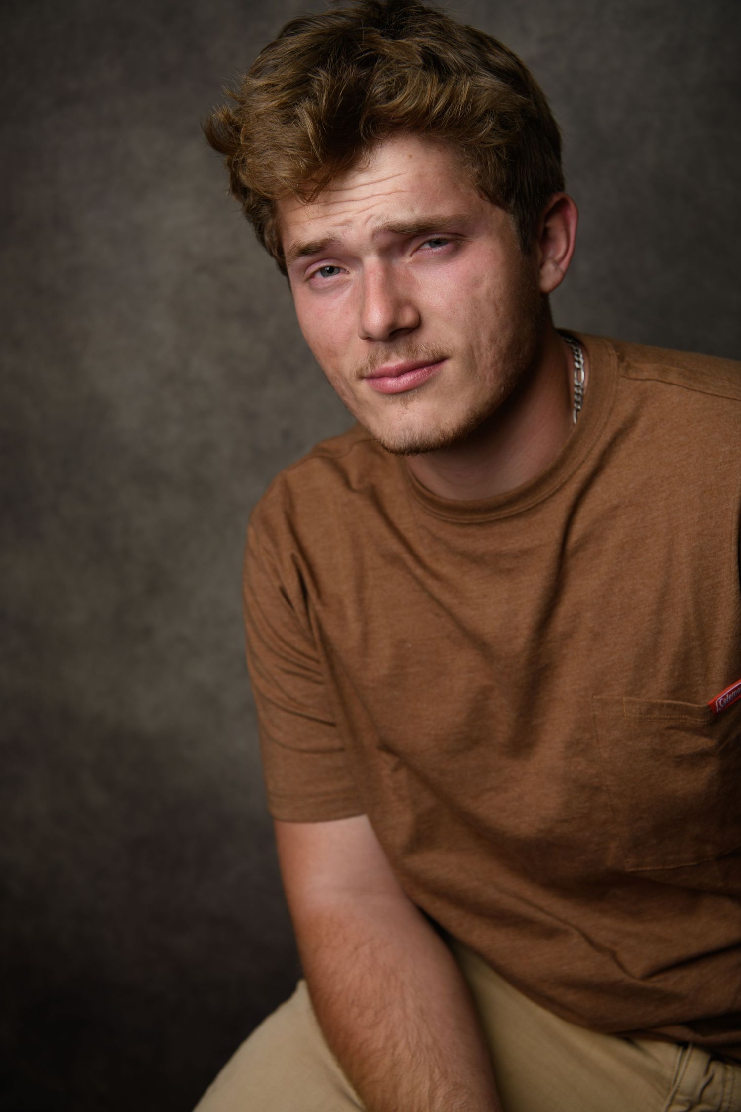 Person in brown t-shirt poses casually against textured dark background.