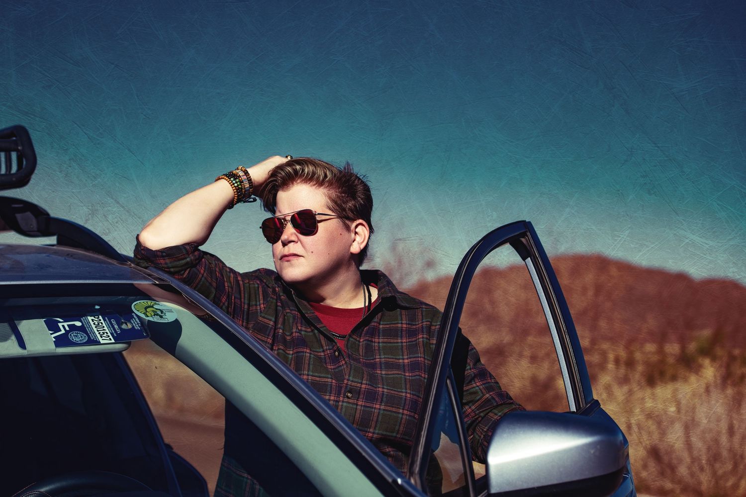 Kelly Kilgore in sunglasses leaning out of car window against desert landscape.