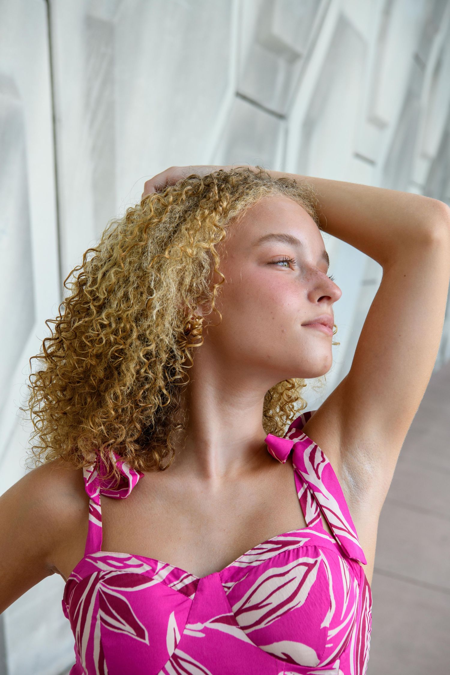 Person in pink floral dress with curly hair posing against white curtains.