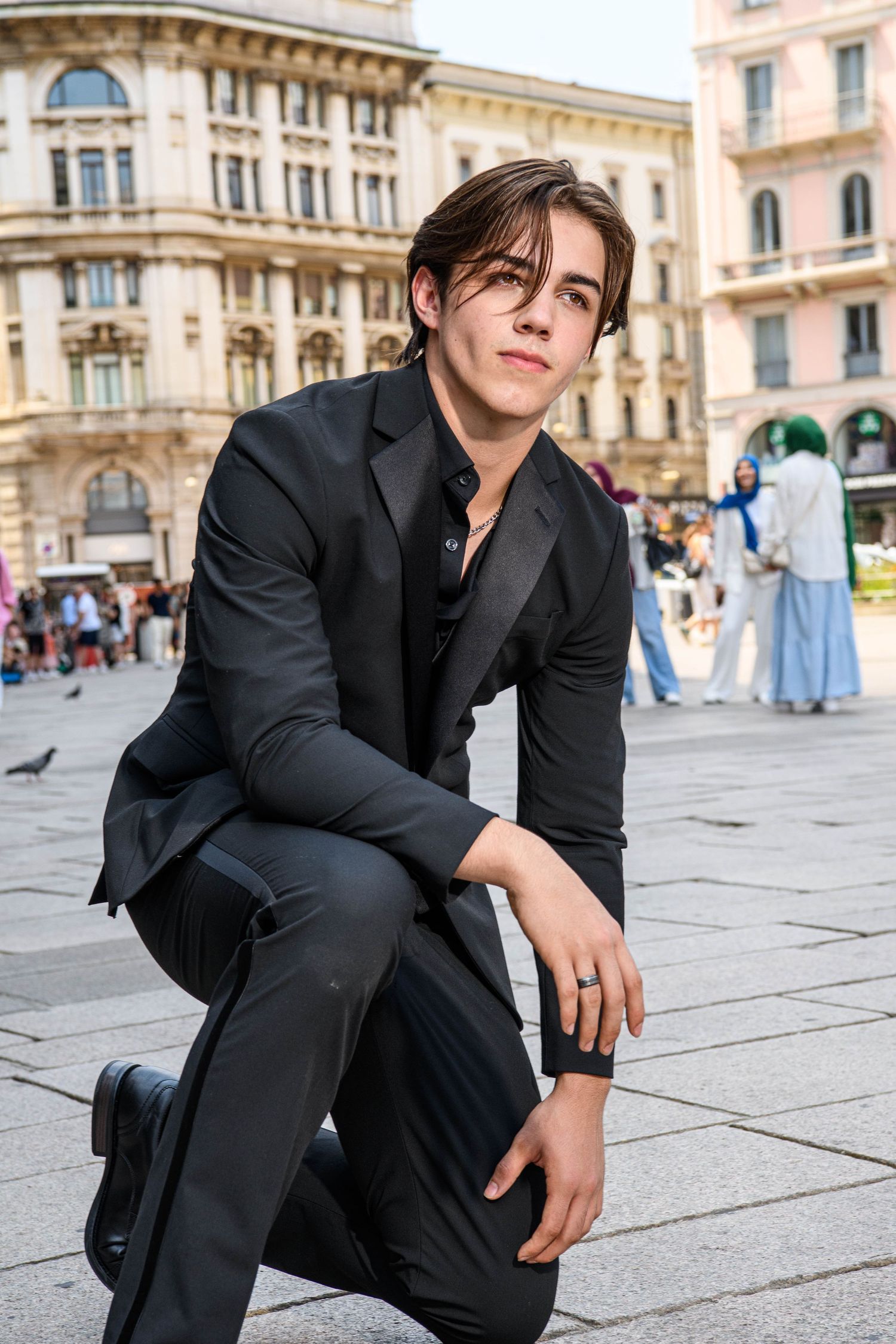 A model in a black suit poses casually while sitting on stone pavement in a historic European city square.