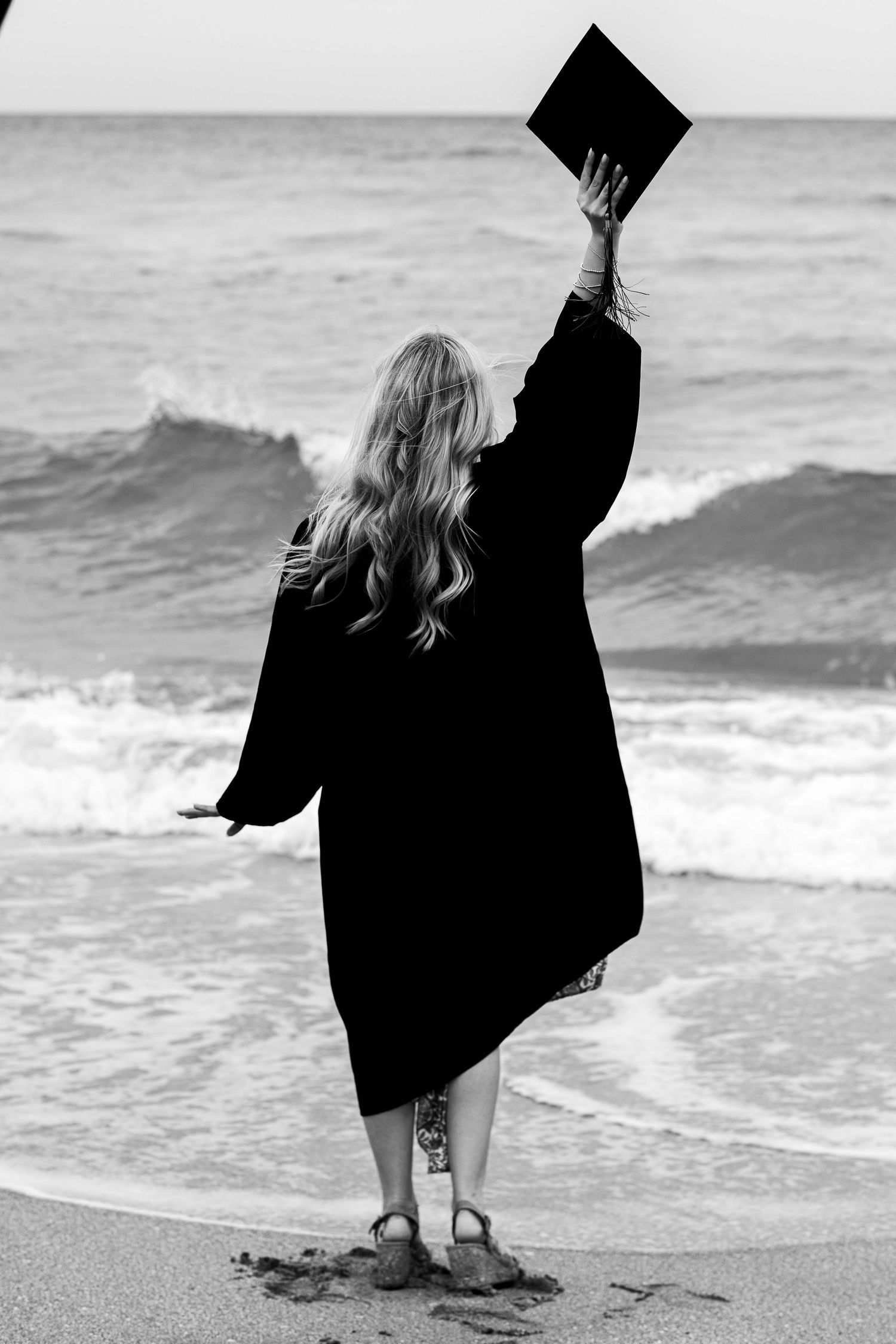 Graduate in black cap and gown raises diploma triumphantly on beach at sunset.