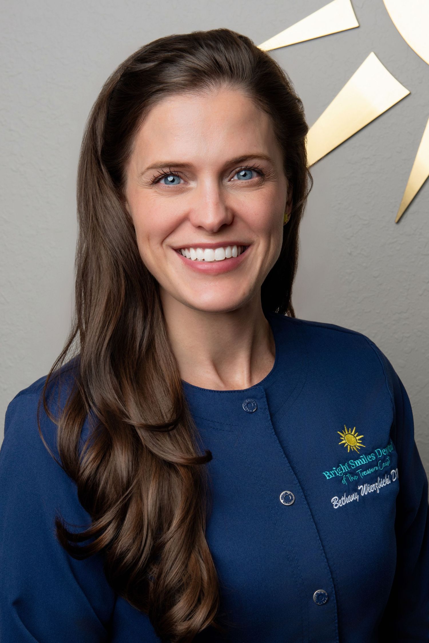 Professional headshot of dental staff member in navy uniform against grey background with gold accents.
