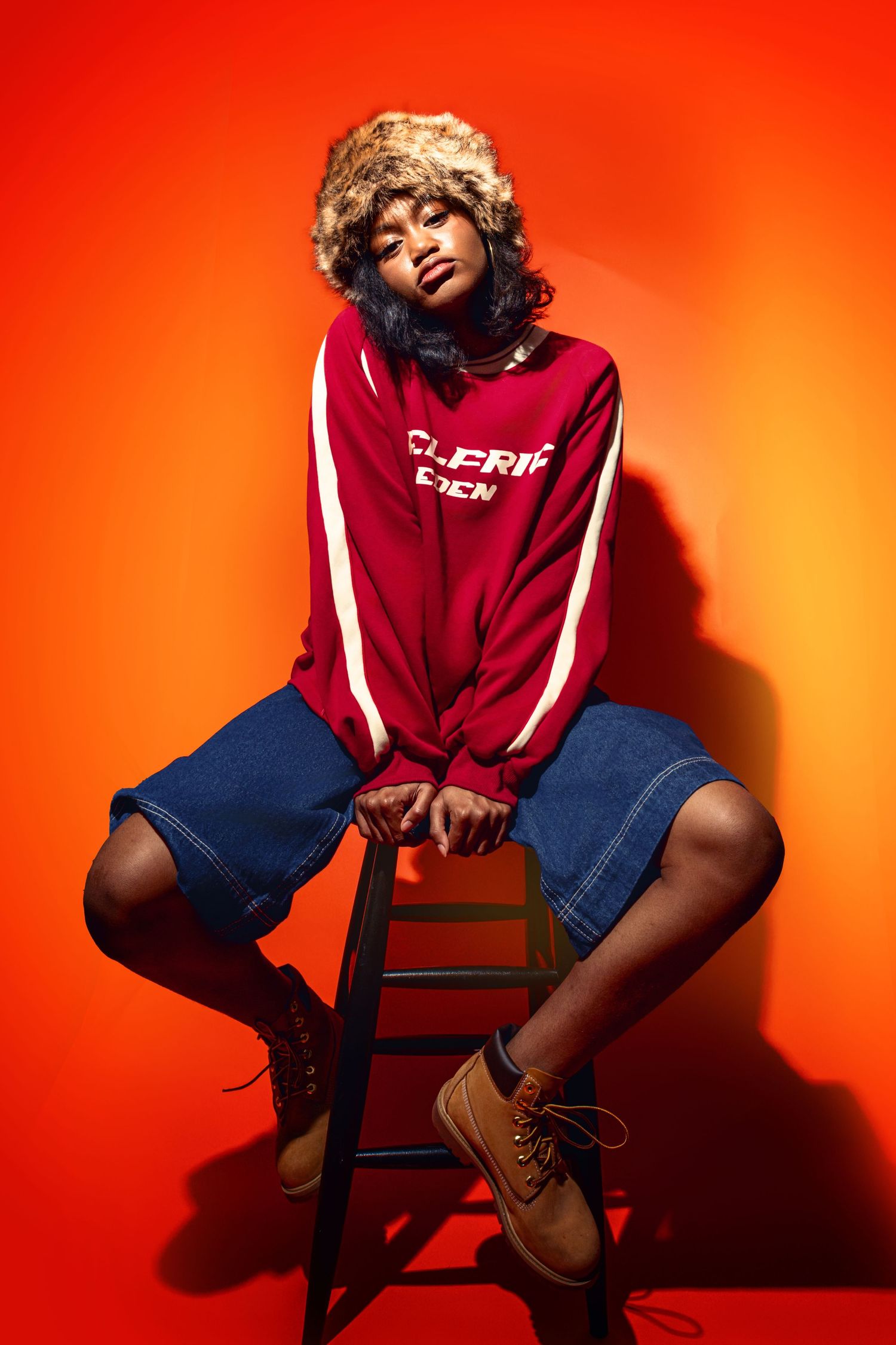 Model in red sweatshirt and denim shorts poses on stool against vibrant orange background.