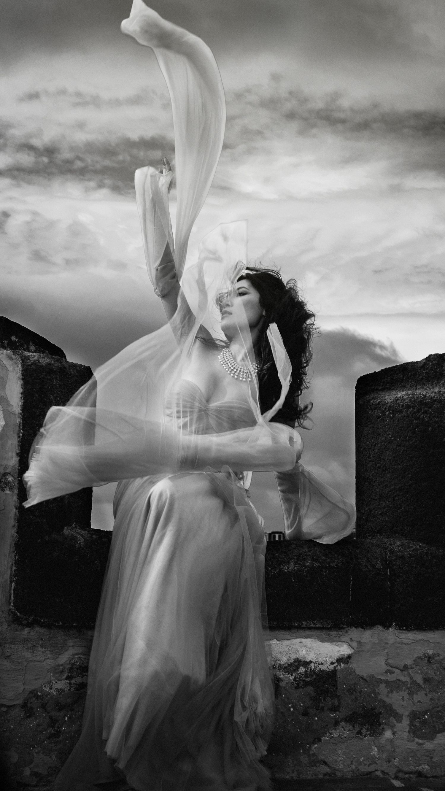 Ethereal black and white image with flowing fabric against a cloudy sky and stone wall.