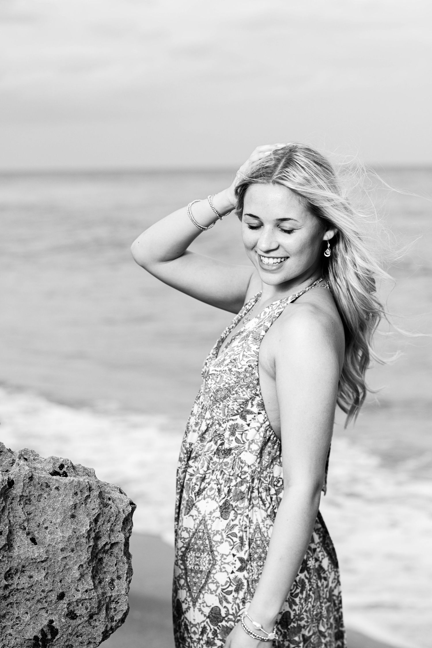 Black and white portrait of woman in floral dress posing against ocean backdrop.