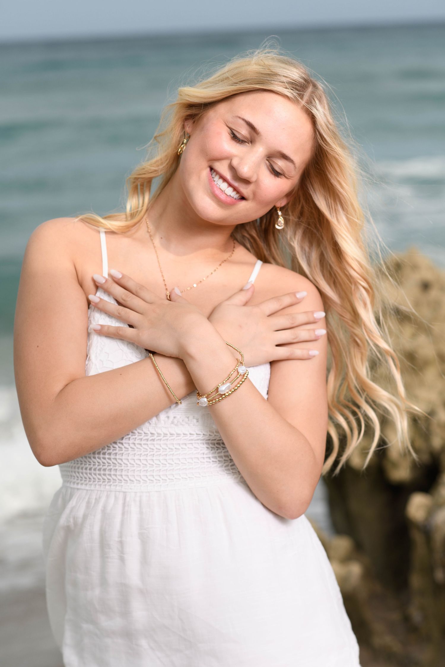 A woman in a white summer dress poses joyfully with closed eyes at the beach.