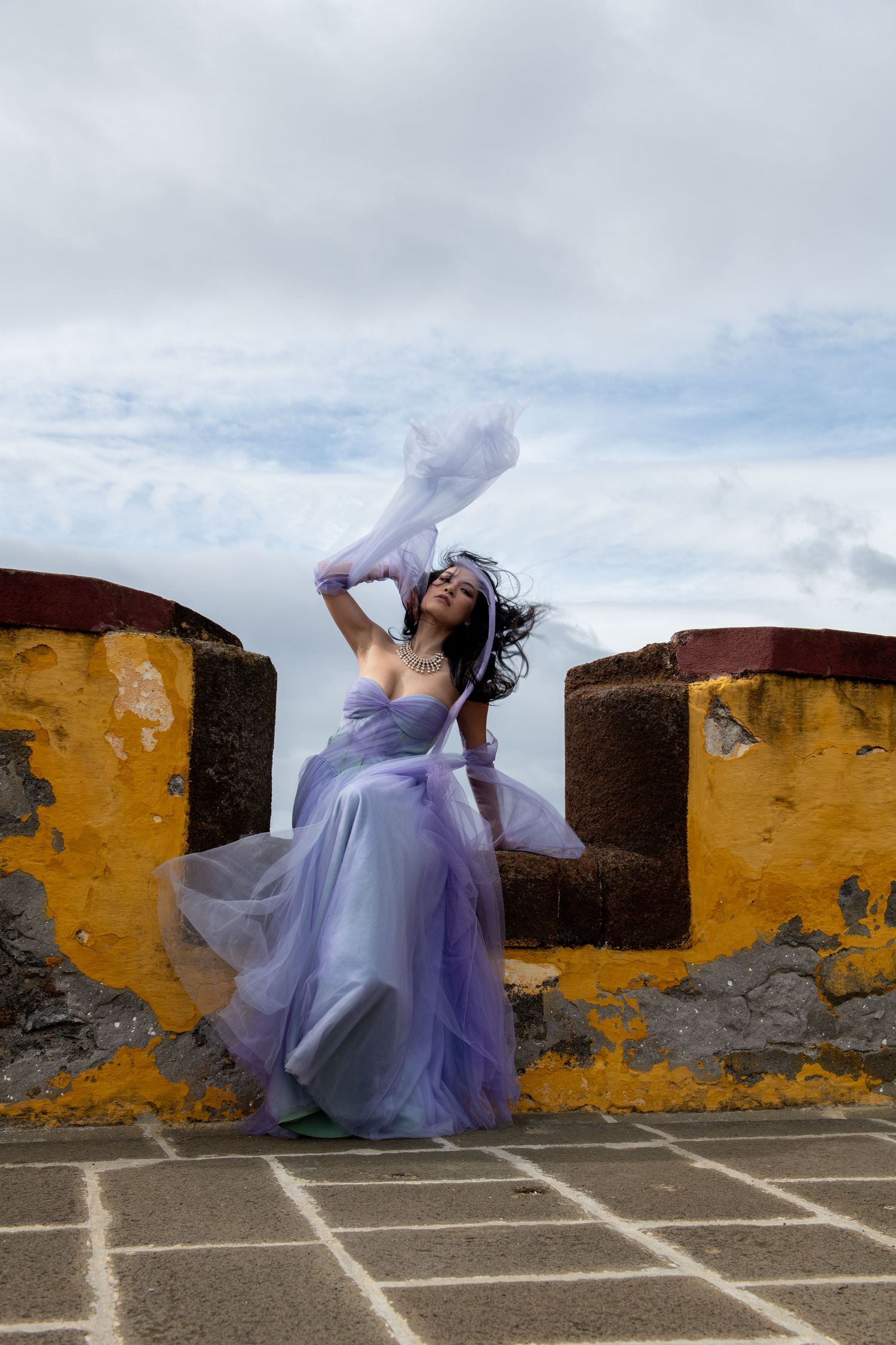 Dancer in flowing purple dress performs dynamic movements against yellow wall outdoors.