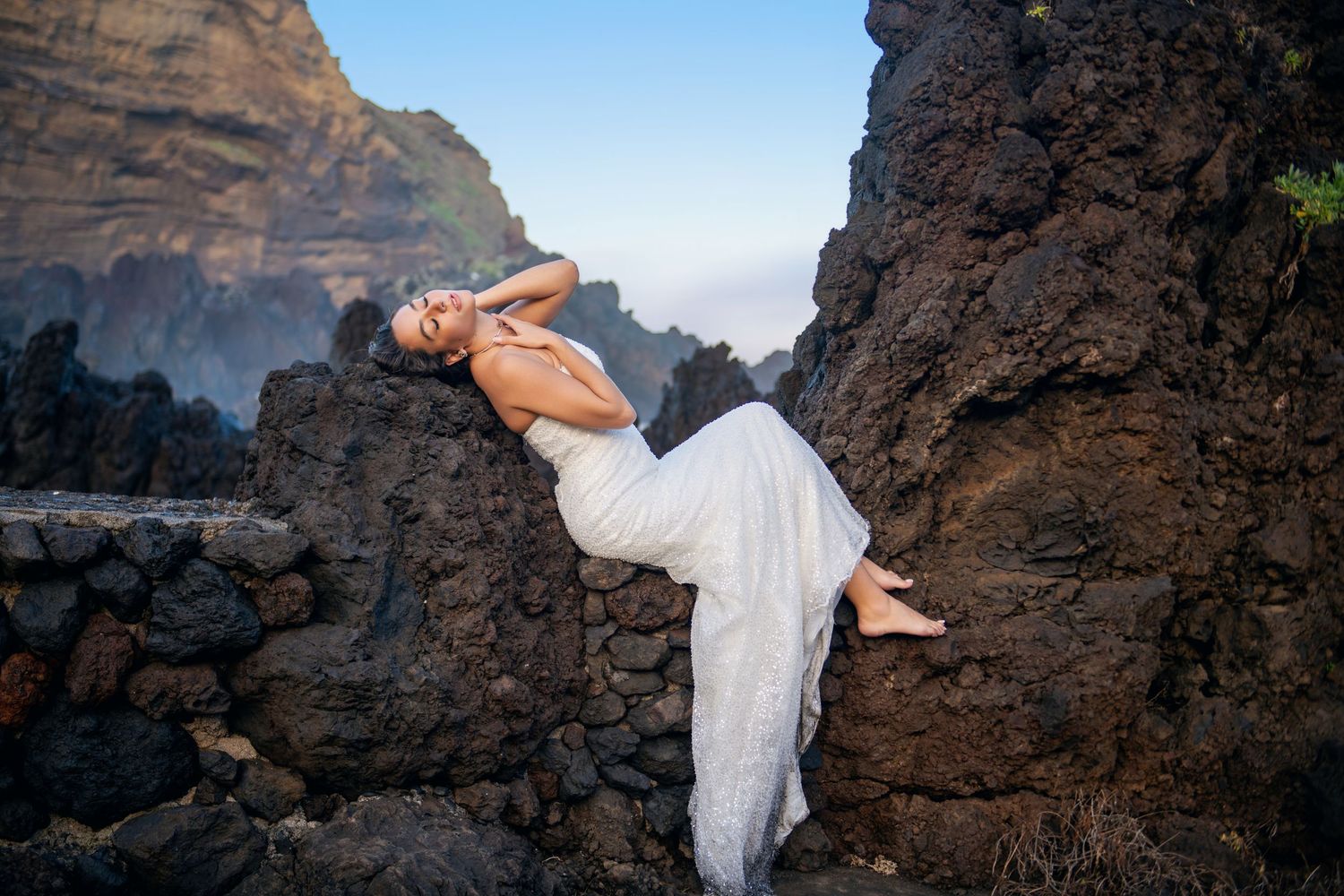 A figure in white fabric poses gracefully on rugged coastal rocks during golden hour.