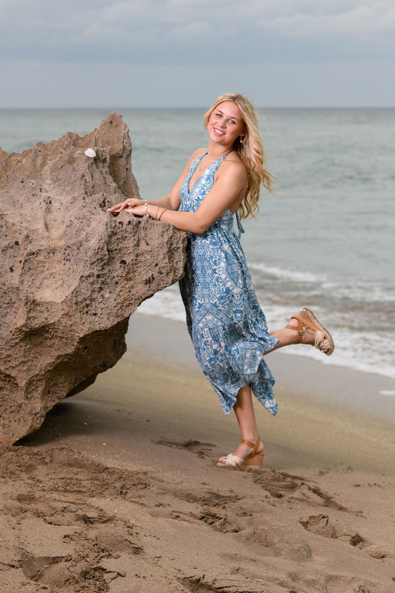 Woman in blue floral dress poses playfully near large rock formations on a beach during sunset.