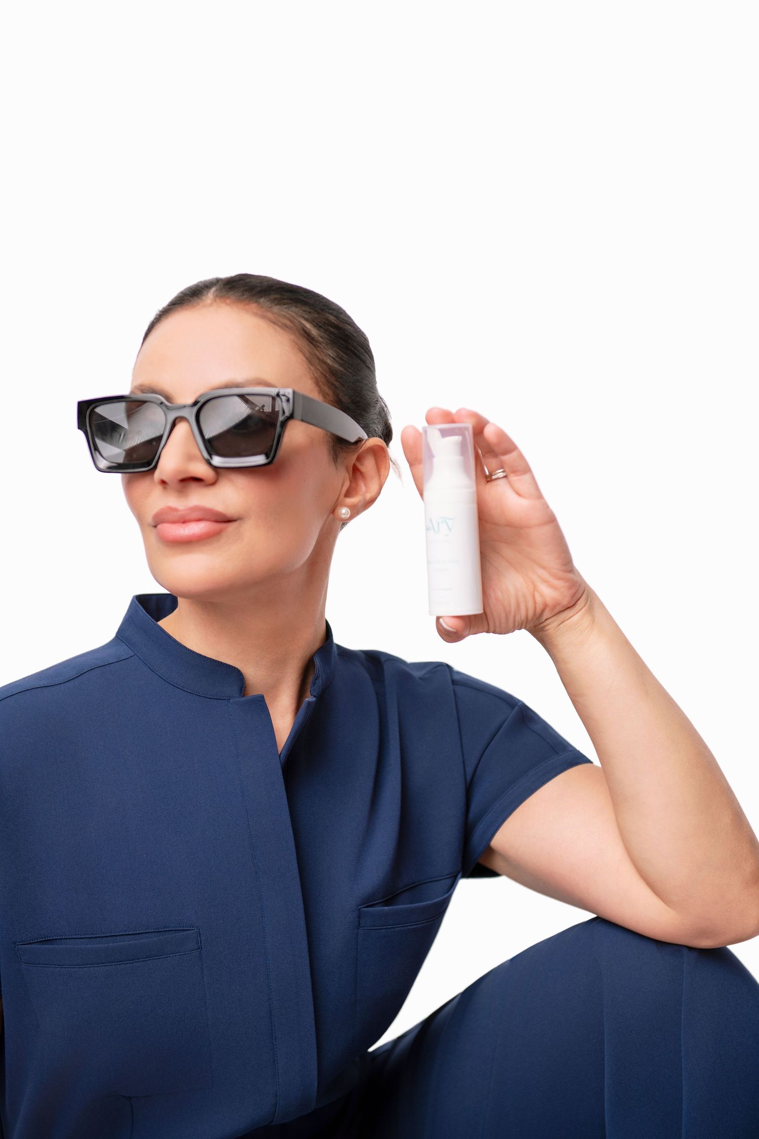 Model in navy outfit displaying white product container while wearing sunglasses.