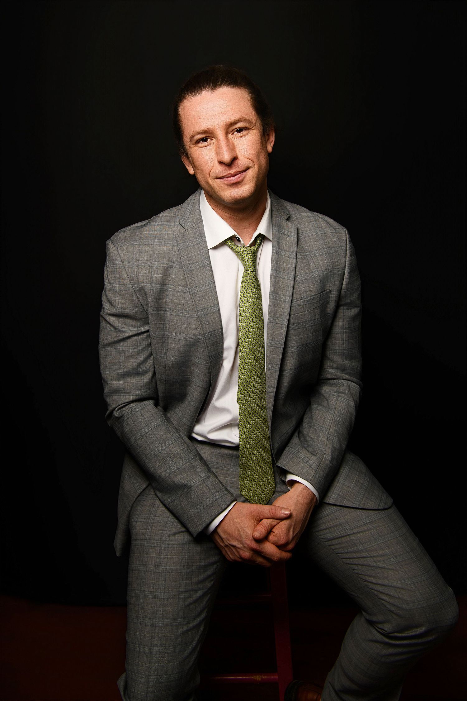 A professional man in a gray suit and green tie sits confidently on a stool against a dark background.