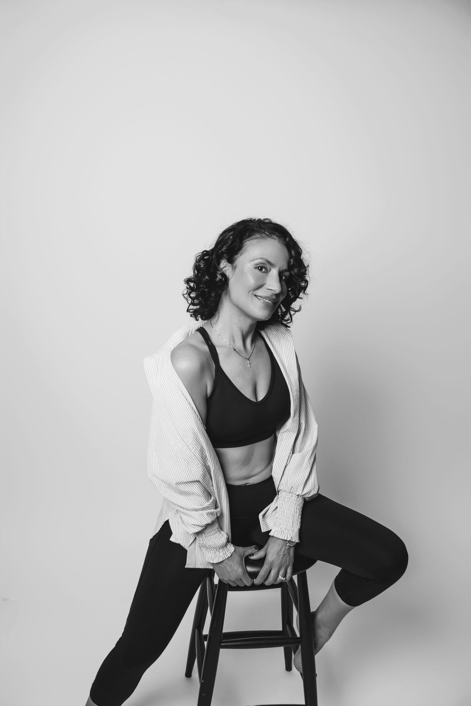 Artistic black and white photo of model in activewear sitting on stool against plain backdrop.