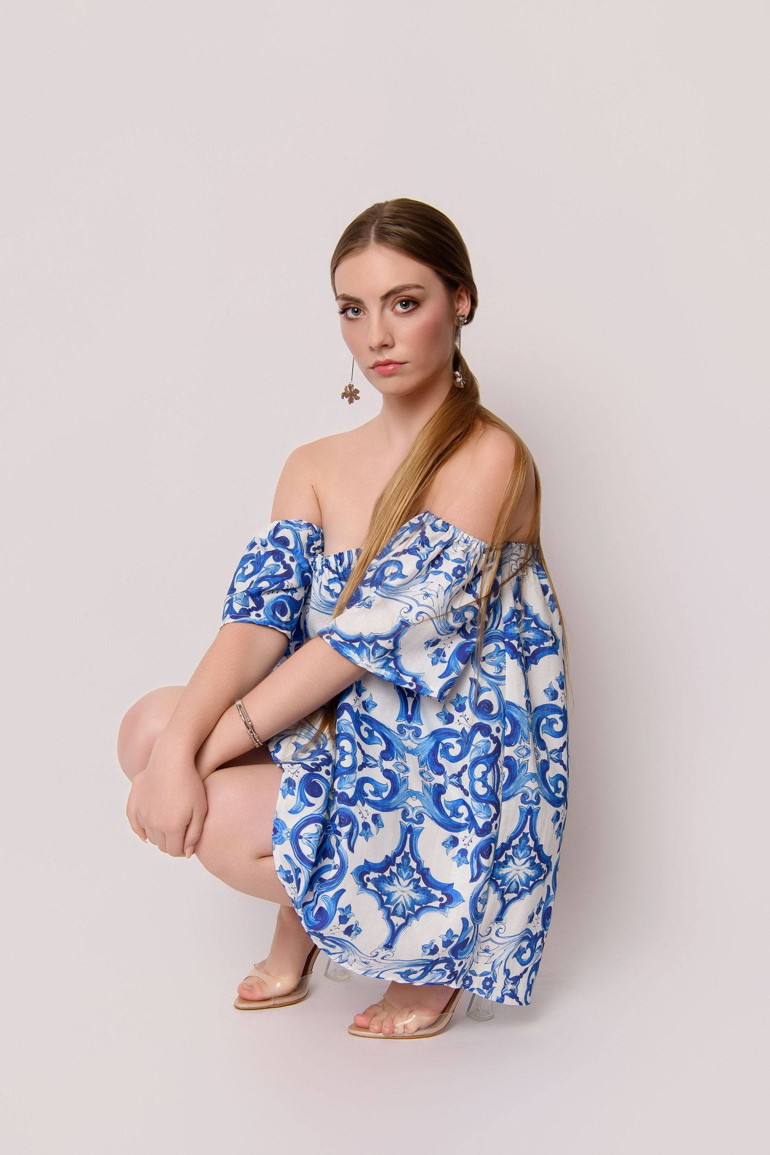 Model in blue and white patterned off-shoulder dress poses against neutral background.