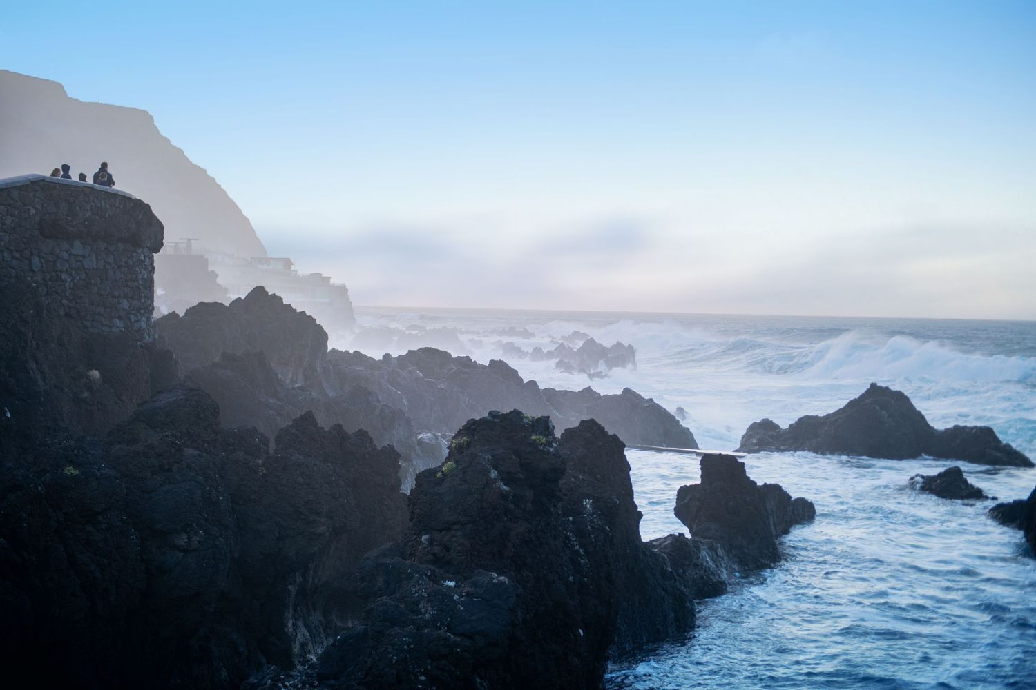 Misty coastal landscape with rocky cliffs and crashing waves against a soft blue sky.