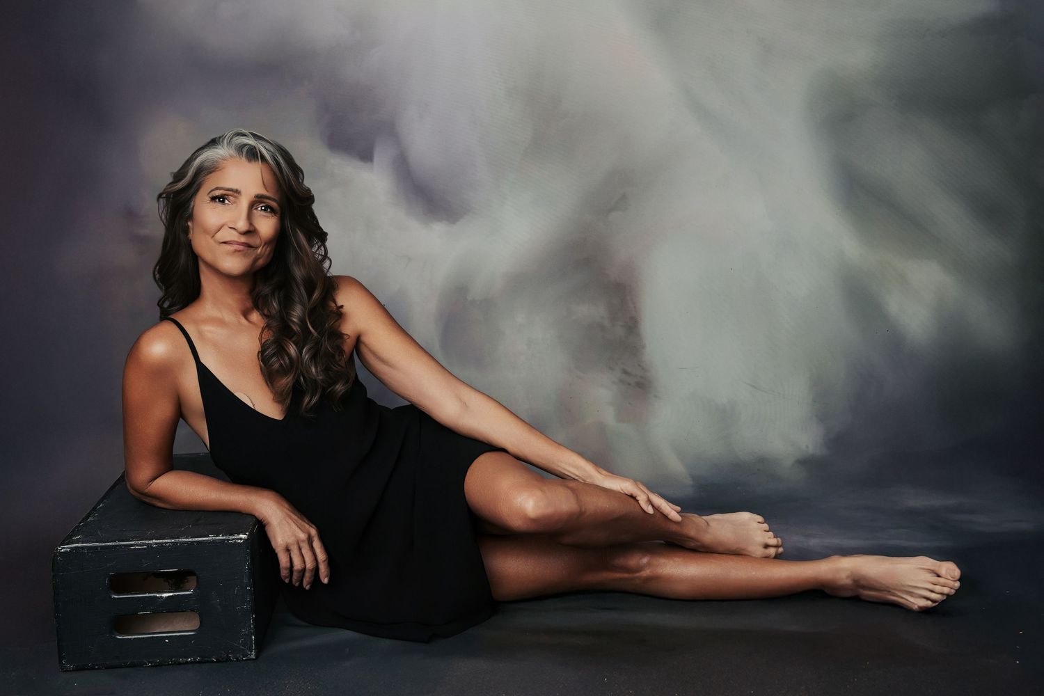 Woman in black dress poses gracefully on floor against abstract painted backdrop, showcasing elegant studio portrait photography.