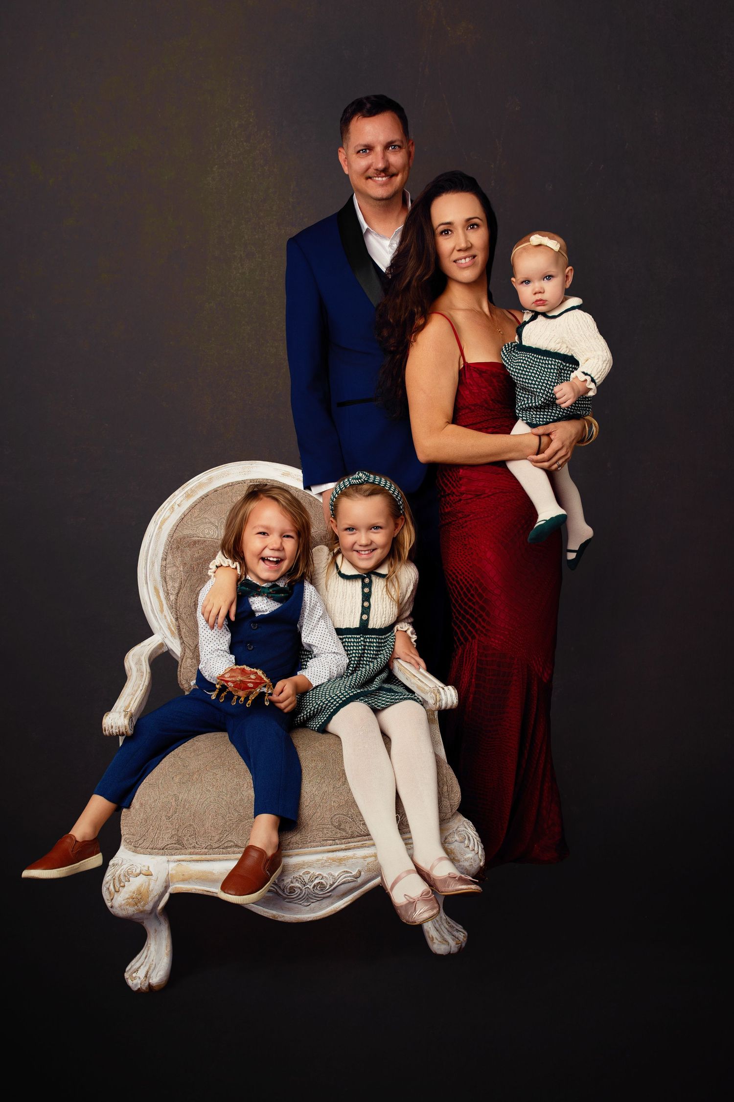 Family portrait series with parents and children sitting on a grey chair against dark background.