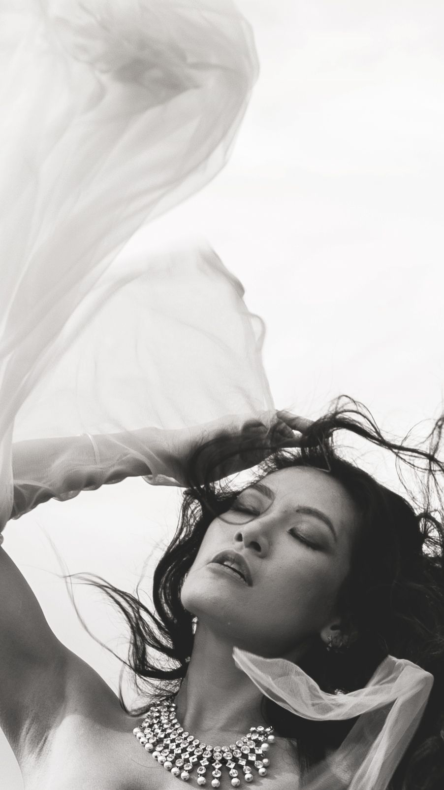 Artistic black and white portrait featuring flowing white fabric and statement necklace against light background.