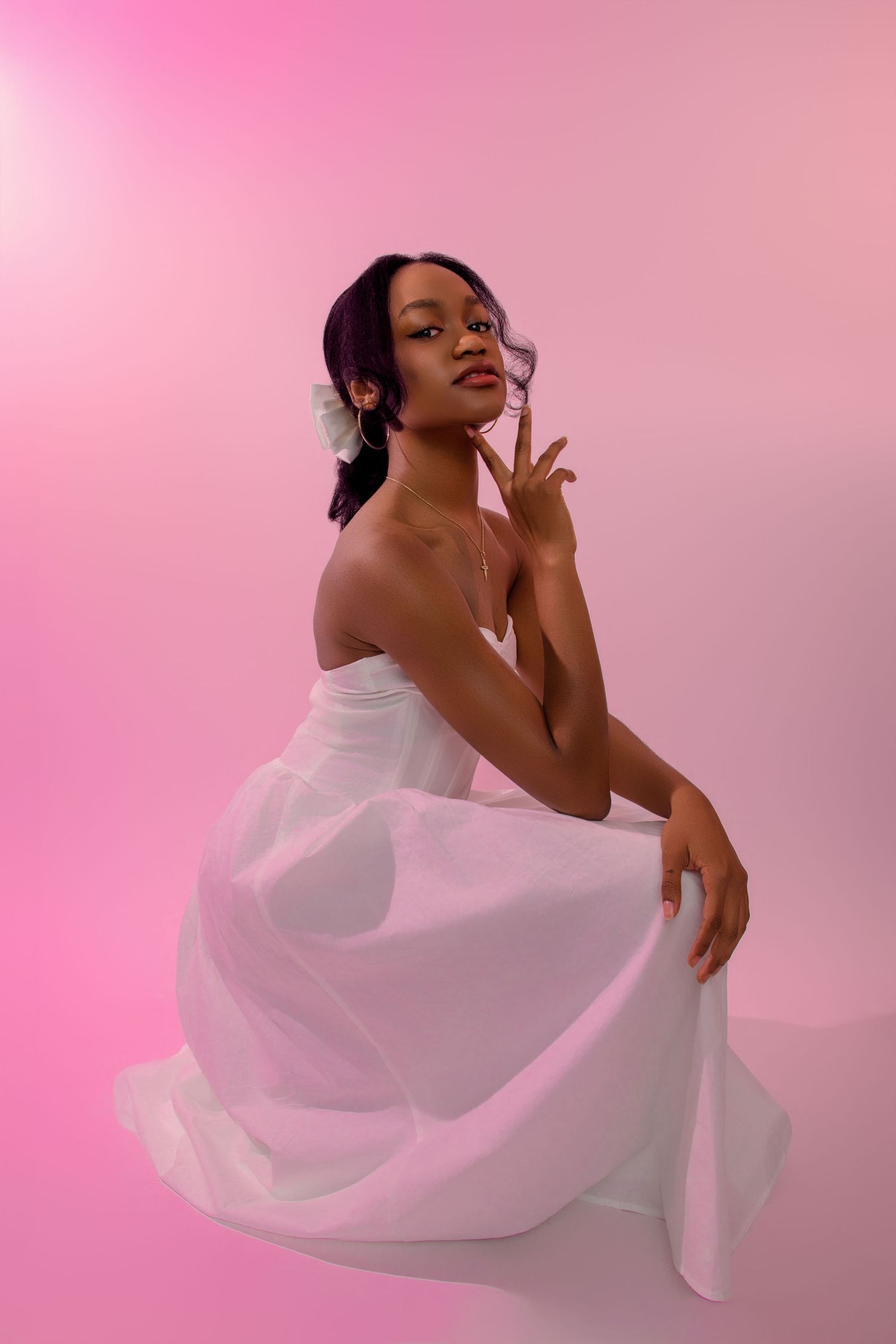 Model in flowing white dress poses gracefully against pink backdrop in artistic photoshoot.