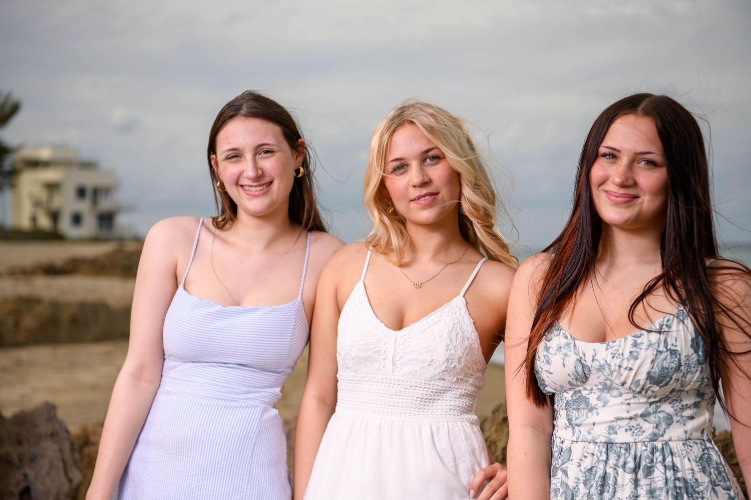 Three people in white and floral dresses pose together outdoors on a sunny day.