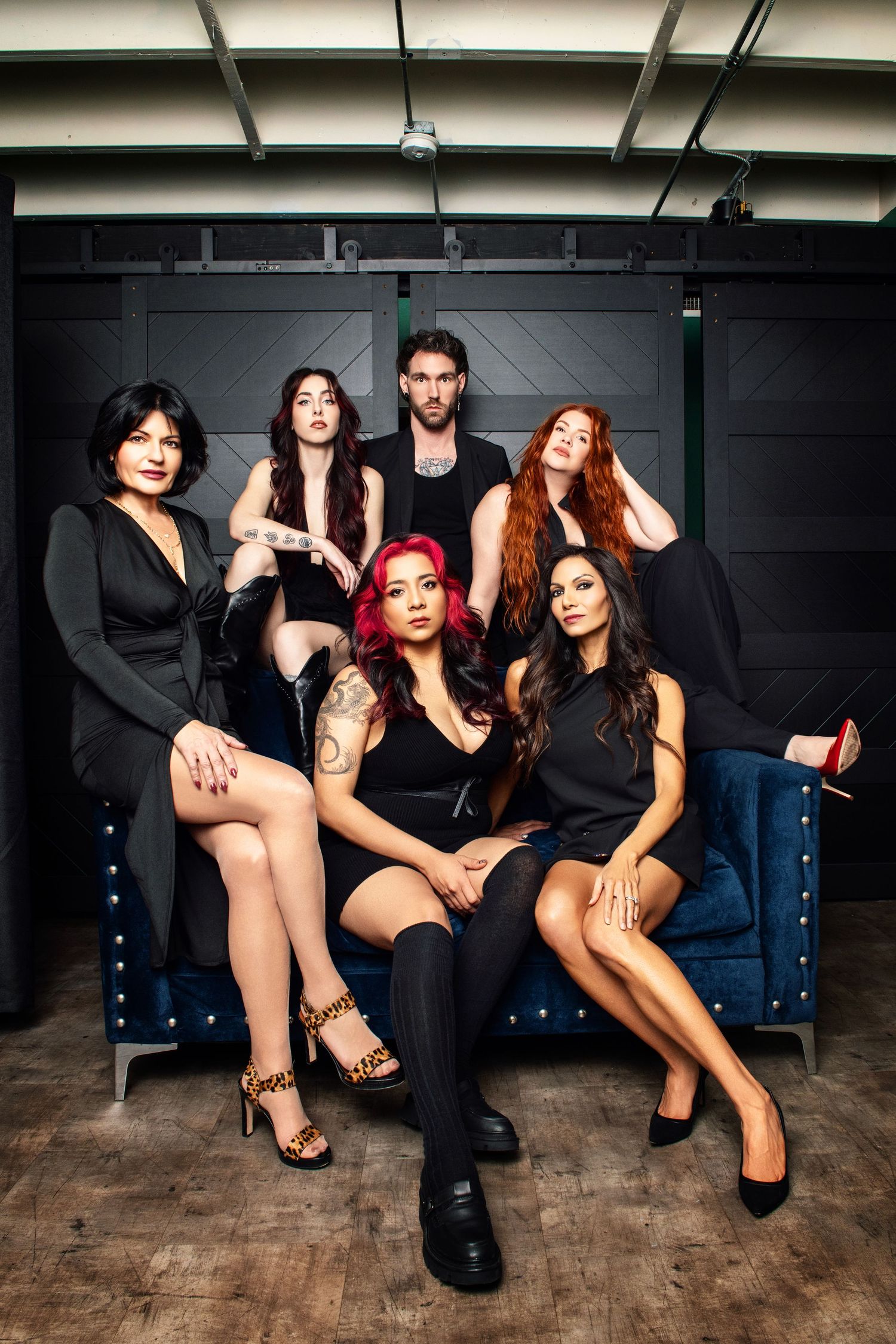 Fashion group photo with people in black attire sitting on blue couch against industrial backdrop.