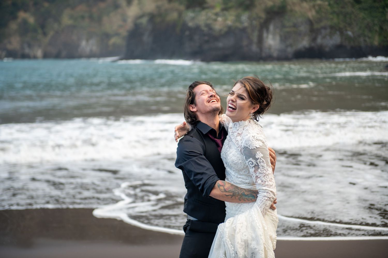 A couple shares intimate moments on a dramatic volcanic beach with waves crashing behind them.