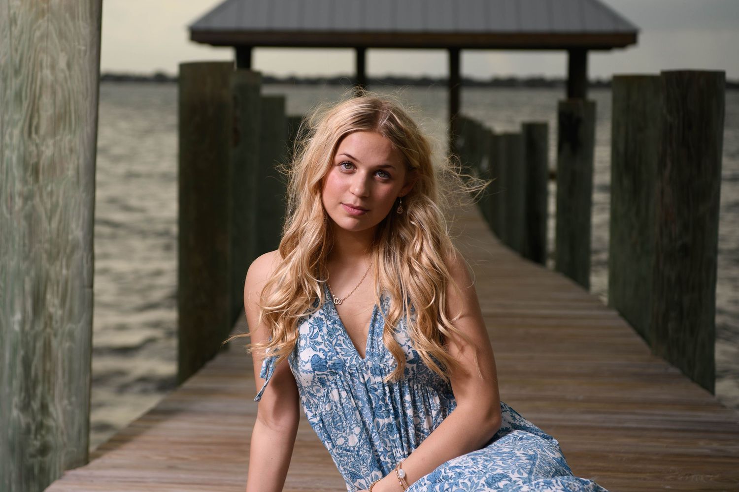 Woman in floral blue dress poses on wooden pier by waterfront at sunset.