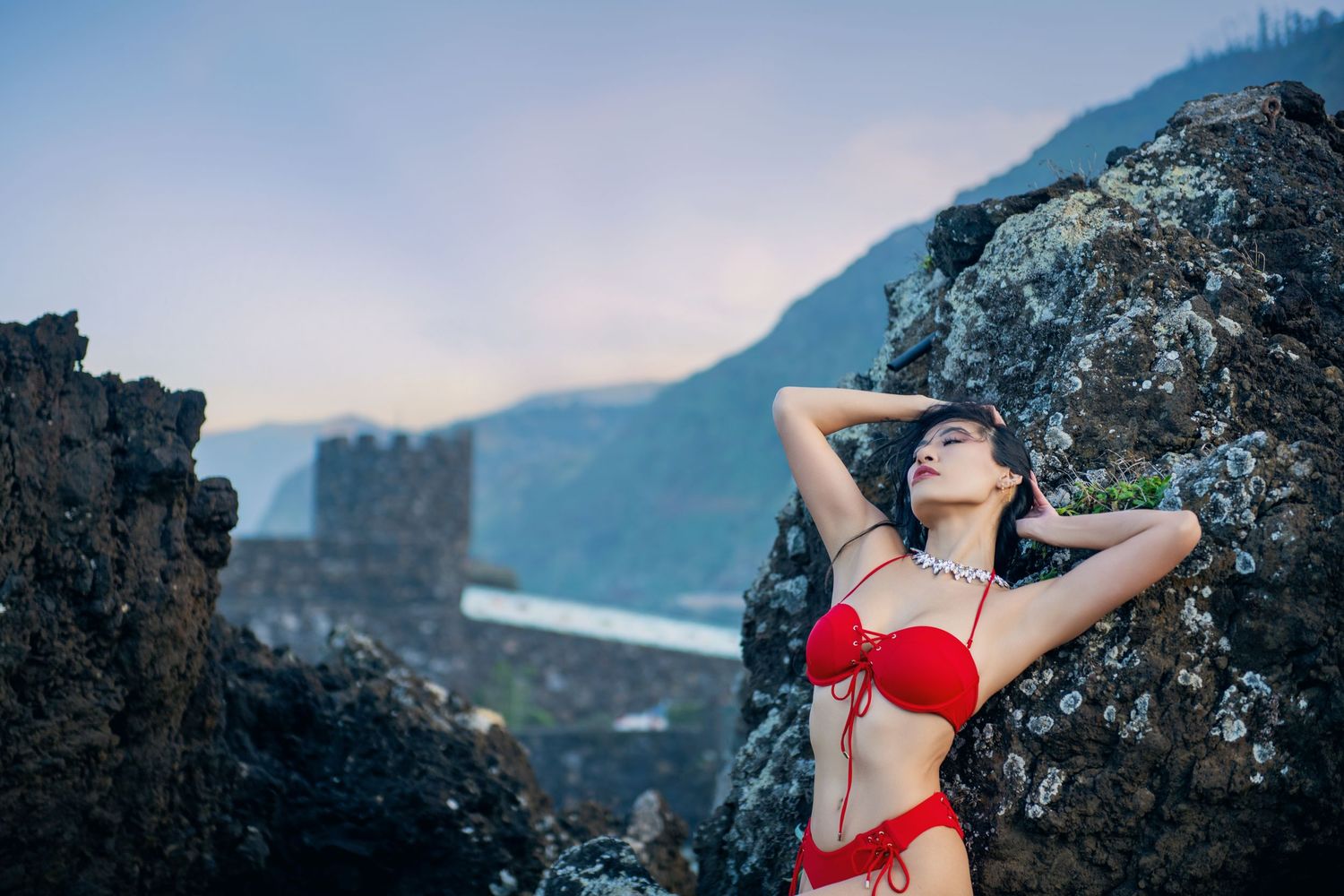 Woman in red swimsuit poses against dramatic rocky coastal backdrop at sunset.
