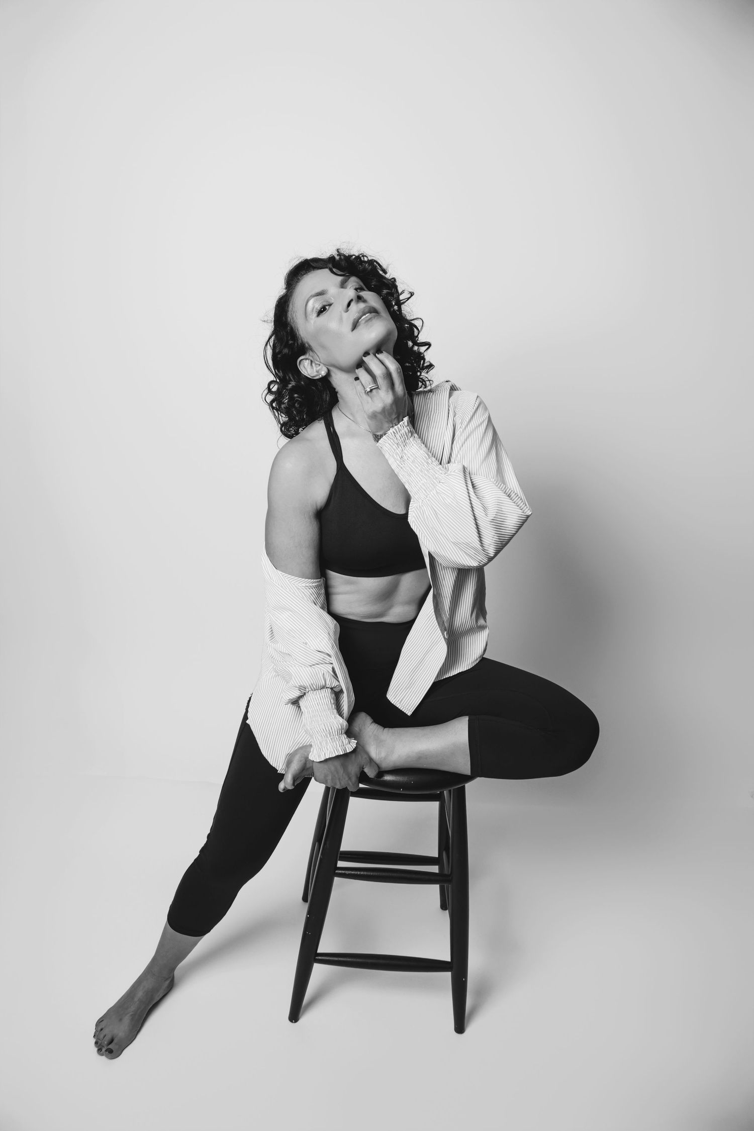 Dramatic pose on wooden stool wearing black athletic wear and white cardigan in monochrome.