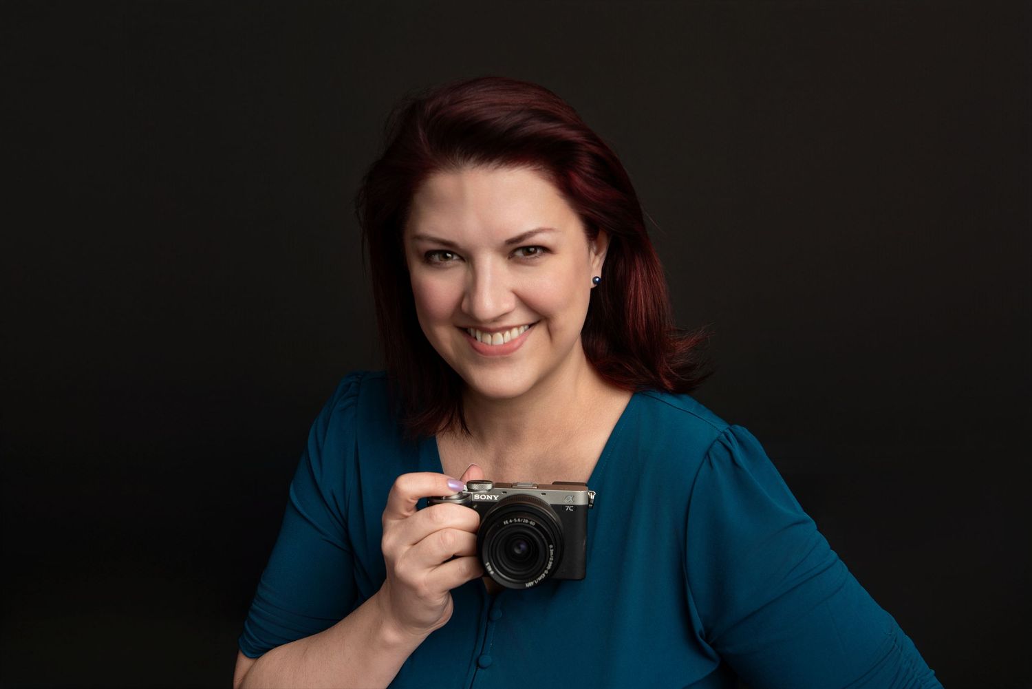 Smiling woman with red hair holding a mirrorless camera, wearing a teal top against a dark background.