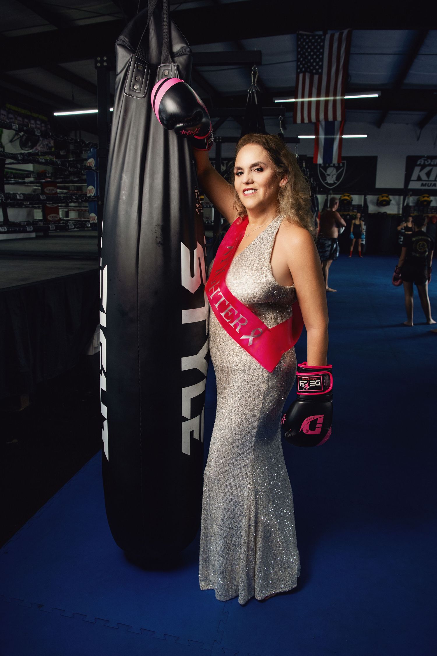 Person in sparkly dress and boxing gloves posing next to punching bag in gym. Promoting Fighting and winning over Breast Cancer