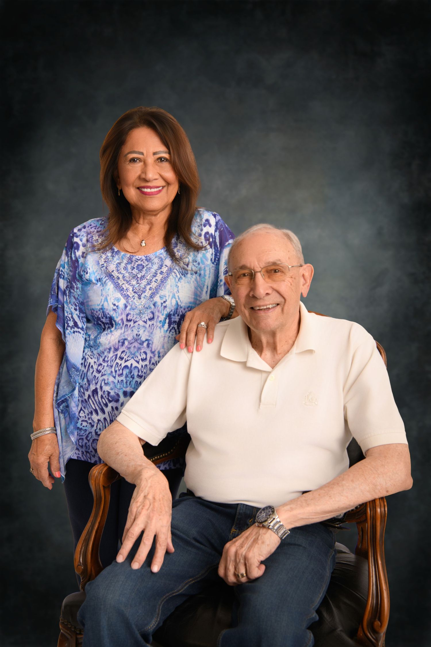 A couple poses together in a wooden chair for a studio portrait against a dark background.