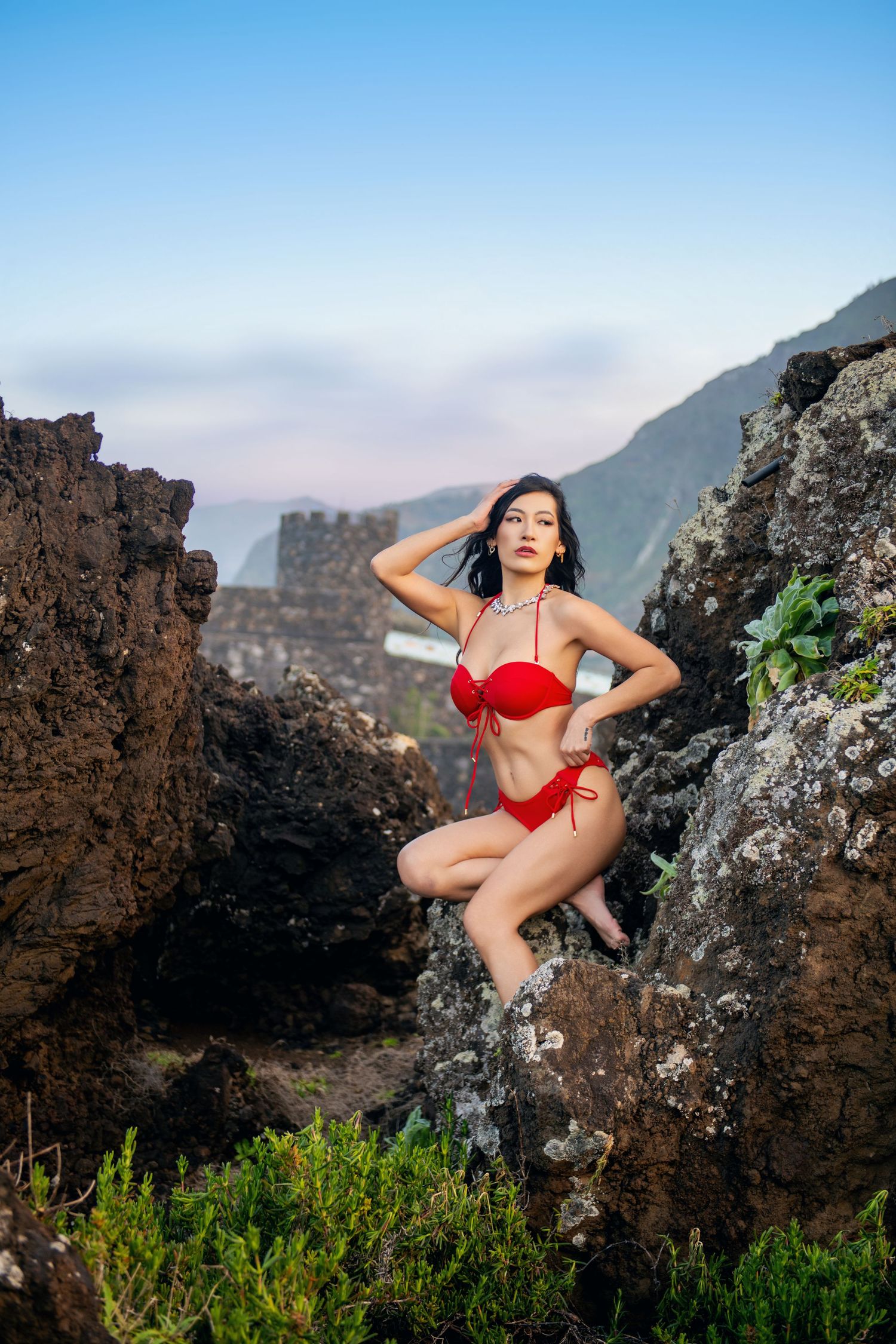 Model in red swimwear poses among rocky coastal formations with mountains in the background.