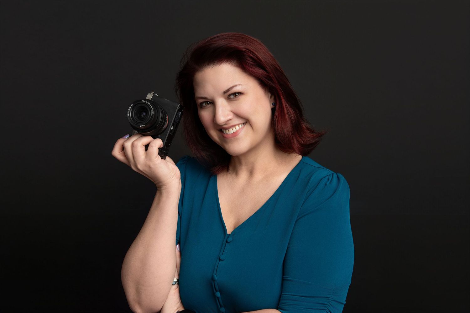 A smiling woman with red hair in a teal blouse holds a professional camera against a dark studio background.